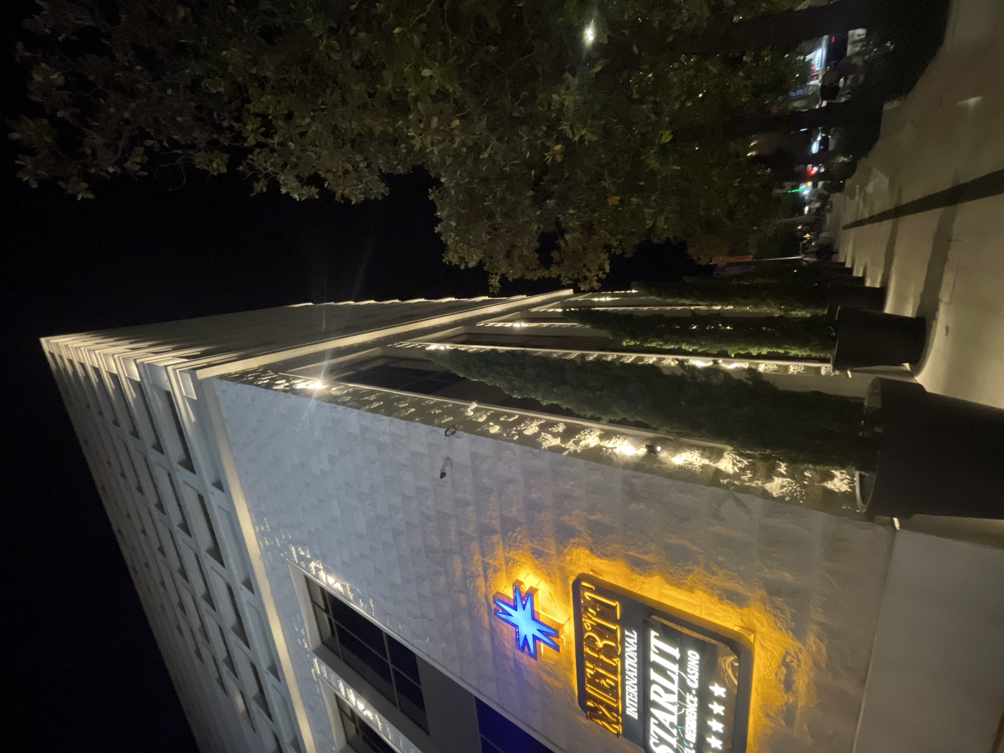 The image shows the Starlit International Residence Casino building at night. The building is tall, with a white facade and many windows. The Starlit logo is visible on the side of the building, lit up with yellow and blue lights. There are trees along the top of the building. The location is in the city of Unknown, Unknown.
