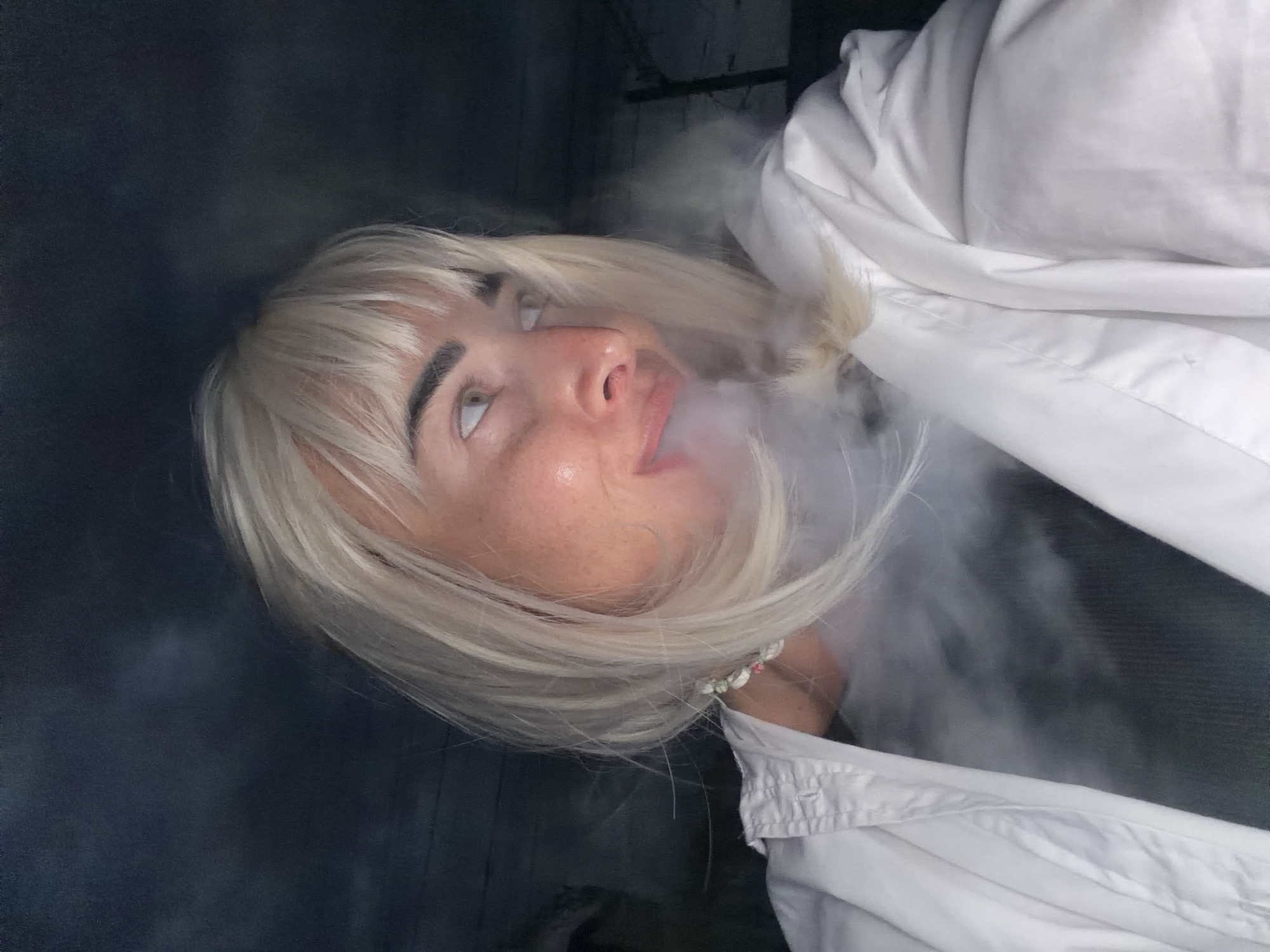 A woman with blonde hair and bangs is exhaling smoke, partially obscuring her face. She is wearing a white collared shirt and a necklace with colorful beads. The background is dark and out of focus, appearing to be an indoor or enclosed space with visible textures that suggest it could be a rustic or industrial setting. There are no identifiable landmarks to suggest the specific location of Sochi, Russia.