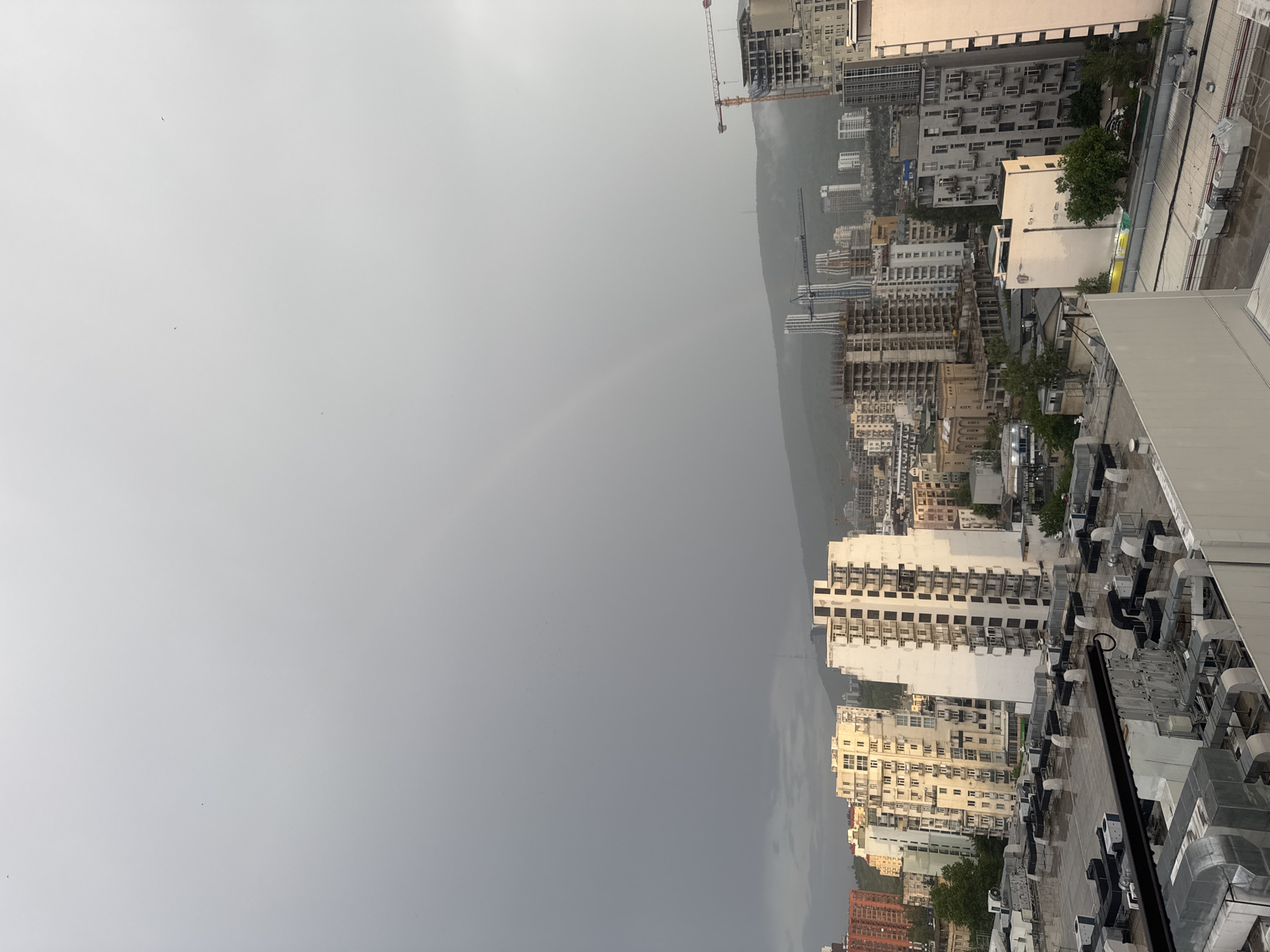 The image shows a rainy cityscape in Tbilisi, Georgia, with a faint rainbow visible in the sky. The view is from a high vantage point, likely a rooftop, looking out over a dense urban area filled with buildings of varying heights and architectural styles. Some buildings are modern high-rises, while others are older, low-rise structures. Construction cranes are visible, indicating ongoing development in the city. The sky is overcast and gray, reflecting the rainy weather conditions.