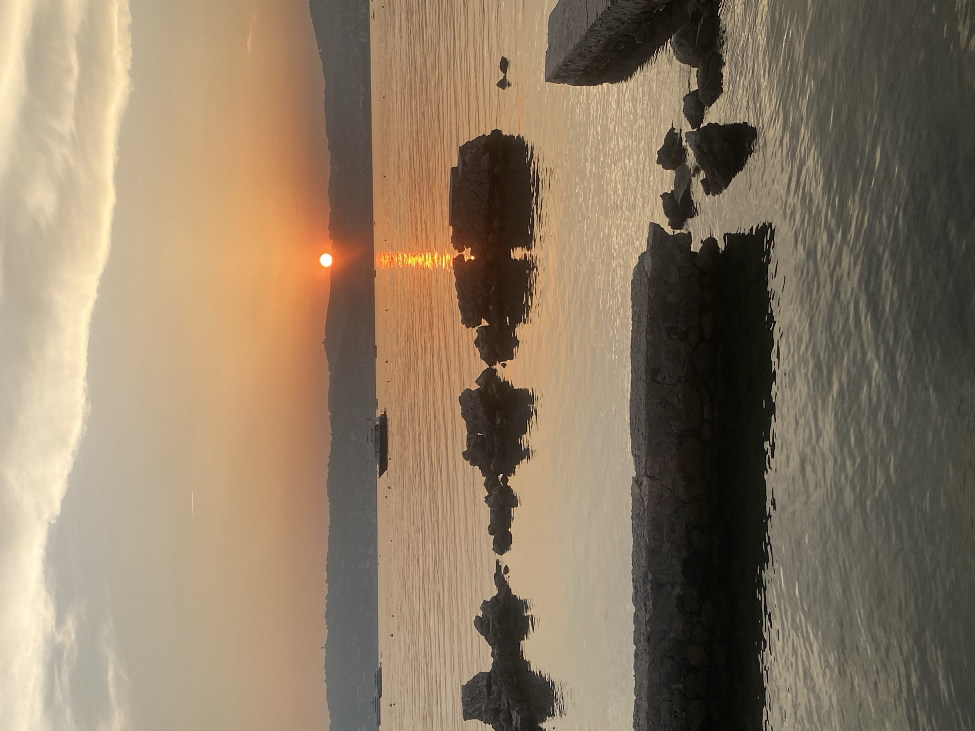 An image captured at sunset over the sea in Antibes, France, showcases a serene waterfront scene. The sun, a vibrant orb of orange, is low on the horizon, casting a warm glow across the sky and reflecting a shimmering path on the water. In the background, hills rise gently, dotted with structures suggesting a town. Several boats, including a dark motor yacht, are moored on the calm sea. In the foreground, weathered stone structures, remnants of a pier or quay, jut out into the water, interspersed with dark rocks. The water itself is calm, with subtle ripples and reflections of the sky and the stone structures. The overall atmosphere is peaceful and tranquil, typical of a Mediterranean coastal evening.