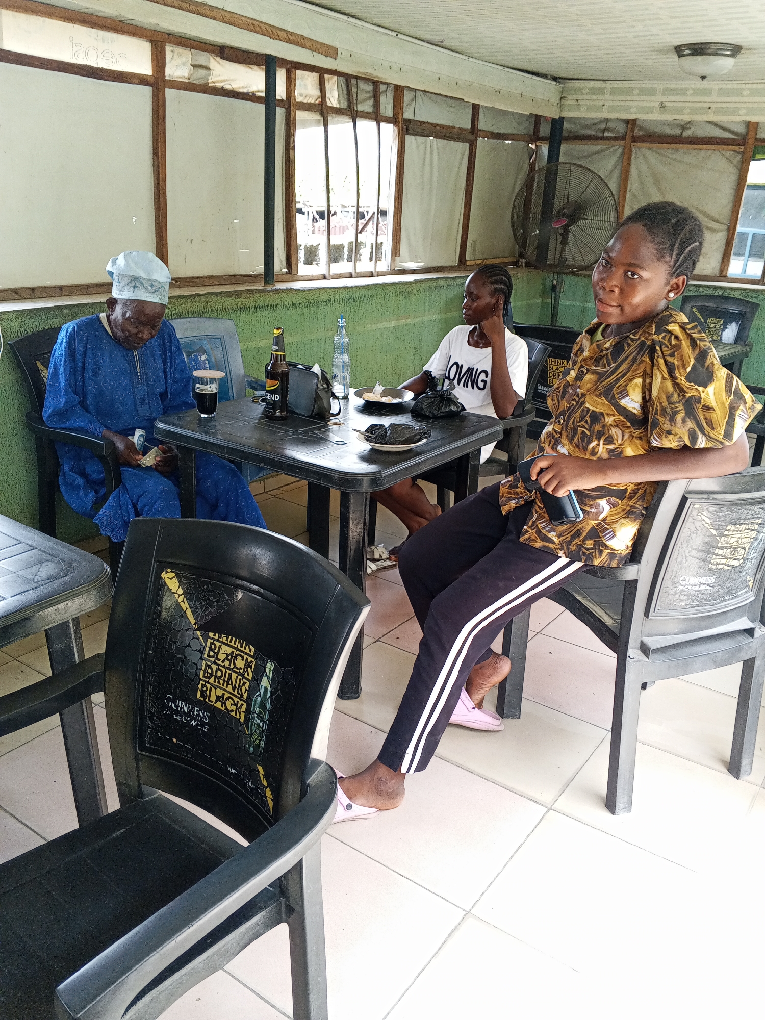 This media file captures a casual indoor scene at what appears to be a local eatery or bar in Zango-ndaji, Nigeria. Three individuals are seated around black plastic tables.
In the foreground to the left, an elderly person, possibly male, is dressed in a blue traditional kaftan and a white cap. They are seated at a table with a glass containing a dark beverage (likely stout) and a bottle of "LEGEND" stout, and appear to be counting or examining money (Naira notes) in their hands.
In the middle background, a younger person in a white t-shirt with the word "LOVING" printed on it is seated, looking towards the left. The table in front of them holds small black plastic bags, possibly containing food.
To the right, a young person in a patterned yellow and brown short-sleeved shirt and black track pants with white stripes is seated, looking directly at the camera with a slight smile. They are holding a mobile phone.
The setting features green and white painted walls, a white tiled floor, and wooden-framed windows with white coverings. A ceiling fan is visible in the background, suggesting a warm climate. Several black plastic chairs, some bearing "GUINNESS" branding and the text "BLACK DRINK BLACK," are arranged around the tables. A bottle of water is also on the table. The atmosphere is relaxed, indicating a social gathering or a break time.