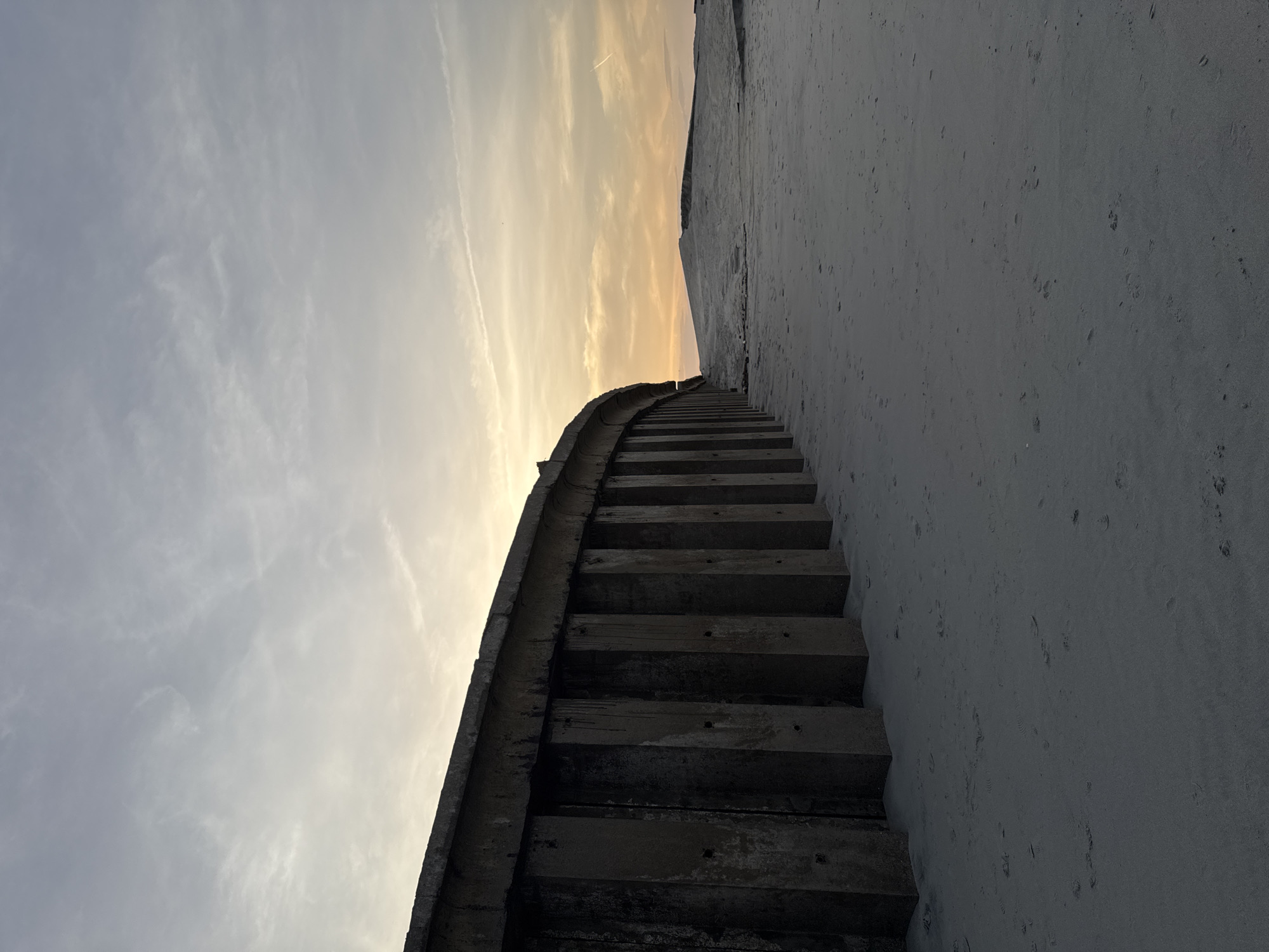A low-angle shot depicts a concrete staircase ascending a steep, sandy slope under a sky with golden and grey hues suggestive of dawn or dusk. The staircase, constructed of dark, weathered concrete steps, curves upwards along the edge of the slope. The adjacent slope is composed of light grey sand. The sky shows a mix of pale grey clouds and a band of golden light near the horizon. A contrail is faintly visible in the upper portion of the sky. The image is angled, placing the staircase on a diagonal. The location is unspecified.