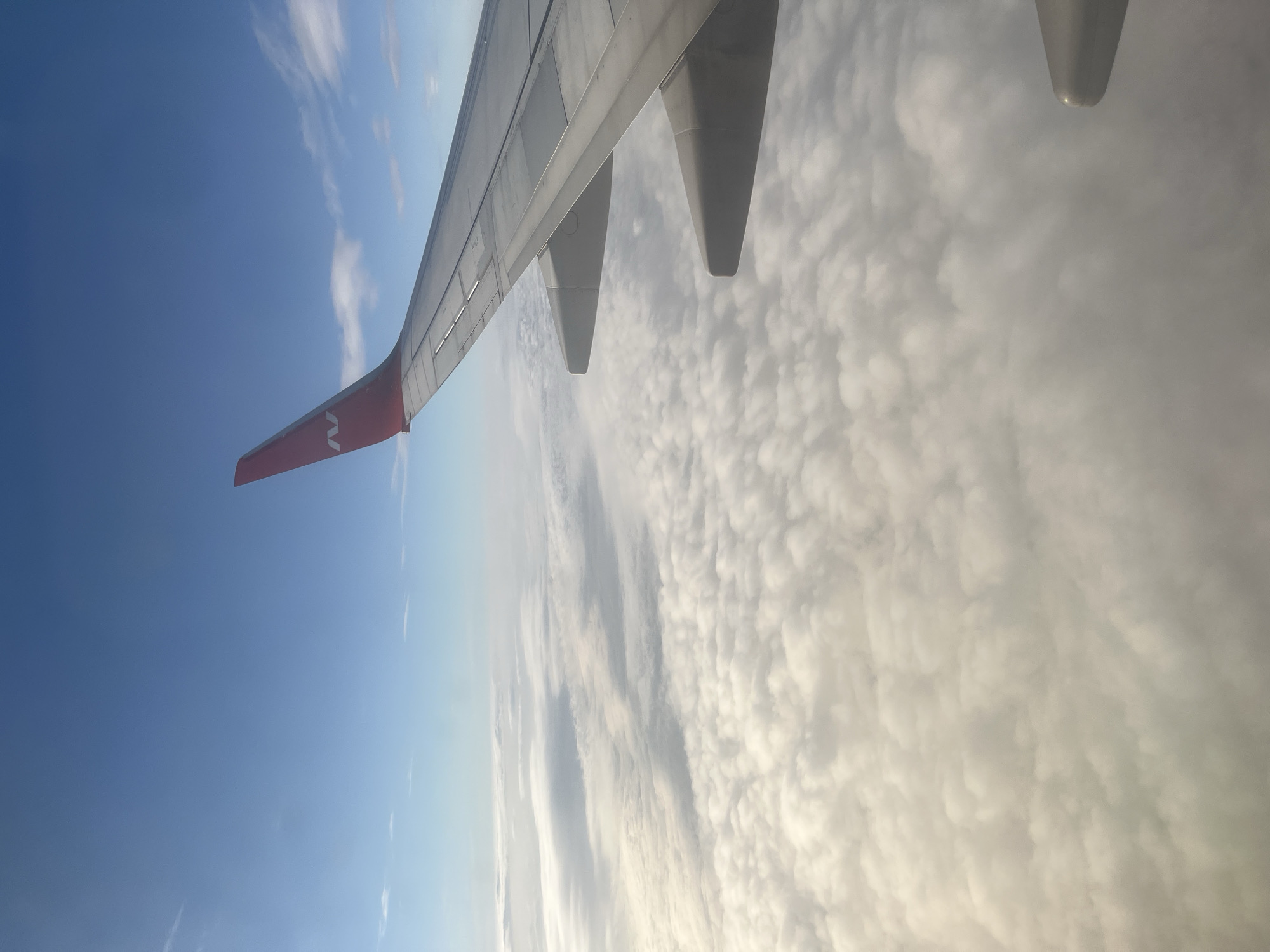 An aerial view from an airplane shows a vast expanse of white clouds below a clear blue sky. The wing of the aircraft, painted with a red tip featuring a white "N" logo, extends into the frame from the right side. The wing is composed of metal panels and has several visible seams and mechanical components. Below the wing, the clouds appear fluffy and layered, with some sections catching the sunlight and appearing brighter than others. The sky above is a gradient of blue, transitioning to a lighter shade near the horizon. The image appears to be taken during daylight hours, with the sun likely positioned out of frame, illuminating the clouds from above. There are no visible people or any indication of the city of Stolbishche, Russia in this image.
