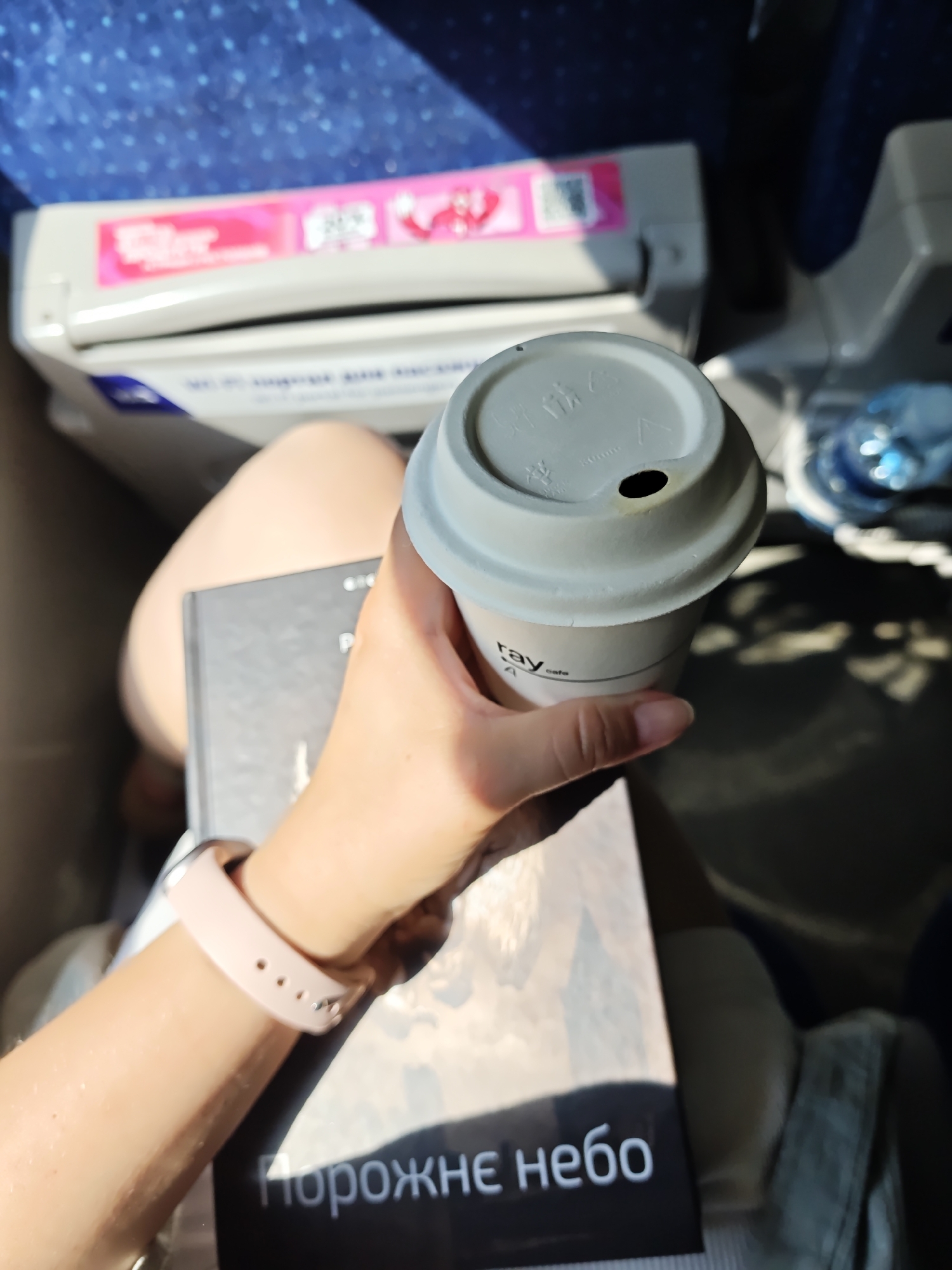 A person is holding a light gray disposable coffee cup with a black lid that has a small opening, and the cup has a logo that reads "ray cafe". The person is also holding a black book with white text that appears to be in Cyrillic. The book has the text "порожнє небо" visible on it. The person is wearing a light pink smartwatch on their left wrist. The background shows a blue patterned surface, likely a seat or upholstery, and a white and pink object, possibly part of a train or bus interior. Sunlight is casting shadows on the surface in the foreground. The scene appears to be indoors, possibly in transit. The location is not identifiable from the image itself.