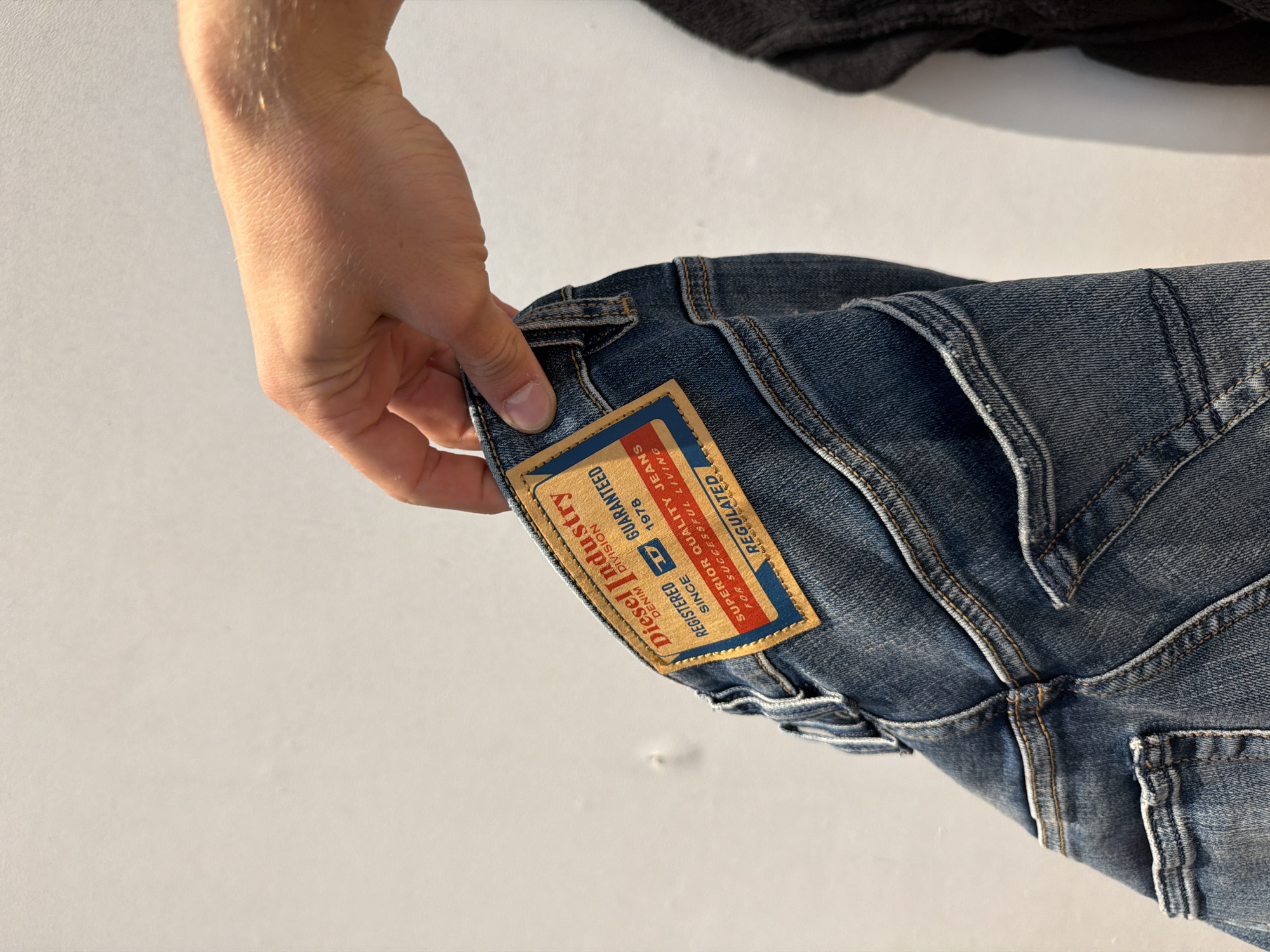 A hand holds up a pair of folded blue denim jeans. The jeans have a Diesel Industry label on the back. The label includes the words "DENIM", "REGISTERED", "GUARANTEED", "SINCE 1978", "SUPERIOR QUALITY JEANS", and "REGULATED". A dark colored garment is visible at the top of the image. The background is a light, solid color.