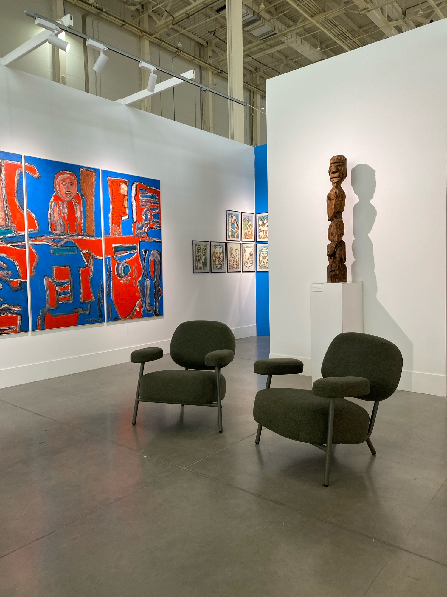 The image depicts an art exhibition space, likely a gallery or museum. Two dark green upholstered armchairs with slender metal legs are positioned in the foreground on a polished concrete floor. To the left, a large triptych painting with bold red and blue abstract figures is displayed on a white wall. In the background, on the right, a tall, dark wooden carved sculpture stands on a white pedestal. Several framed artworks, appearing to be drawings or prints of figures, are mounted on a blue wall section further back. Overhead, track lighting illuminates the space. The environment appears to be a modern, indoor exhibition hall. There are no people visible in the image.