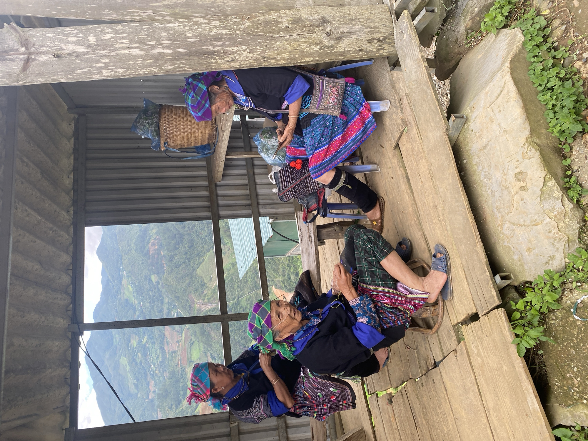 Within an open-sided, corrugated metal and wooden shelter in Dế Xu Phình, Vietnam, three elderly women are observed. The shelter features a corrugated metal roof and rear wall, wooden floorboards, and wooden support posts.
The woman on the left, positioned behind a low wooden bench, wears a dark long-sleeved top with blue accents, a plaid skirt, and a pink and blue plaid headscarf. Her hands are engaged in an activity that appears to be textile-related, possibly braiding or weaving.
The woman in the center sits on a small, round wooden stool. She wears eyeglasses, a dark long-sleeved top with blue-patterned sleeves, plaid green pants, a colorful embroidered apron-like garment, and a green and purple plaid headscarf. Her hands are occupied with a small, dark object, indicating crafting or fine motor work. Her feet are clad in blue open-toed sandals.
The woman on the right sits on a blue plastic stool, leaning against a rough-hewn wooden post. She wears a dark long-sleeved top with blue accents, a vibrant pink and blue patterned skirt, and a purple and blue plaid headscarf. A patterned shoulder bag hangs from her right shoulder. She holds and observes a silver-colored smartphone with both hands. Her left leg features a black fabric wrap or gaiter, and her foot wears a brown open-toed sandal.
On a wooden shelf behind the woman on the right, a woven basket contains a blue plastic bag filled with green, leafy material. Additional bags are visible near her stool.
The shelter overlooks a vast, verdant mountainous landscape under a partly cloudy sky. The mountains are characterized by dense foliage and visible terraced agricultural fields. A utility wire diagonally traverses the upper left portion of the background view. A structure with a green corrugated roof is visible on a distant slope, and a narrow waterfall or cascade descends down a mountain face in the lower right background.
The wooden floor of the shelter exhibits wear, with visible gaps between planks and metal support beams beneath. The ground surrounding the shelter consists of exposed soil, rocks, and patches of green vegetation.