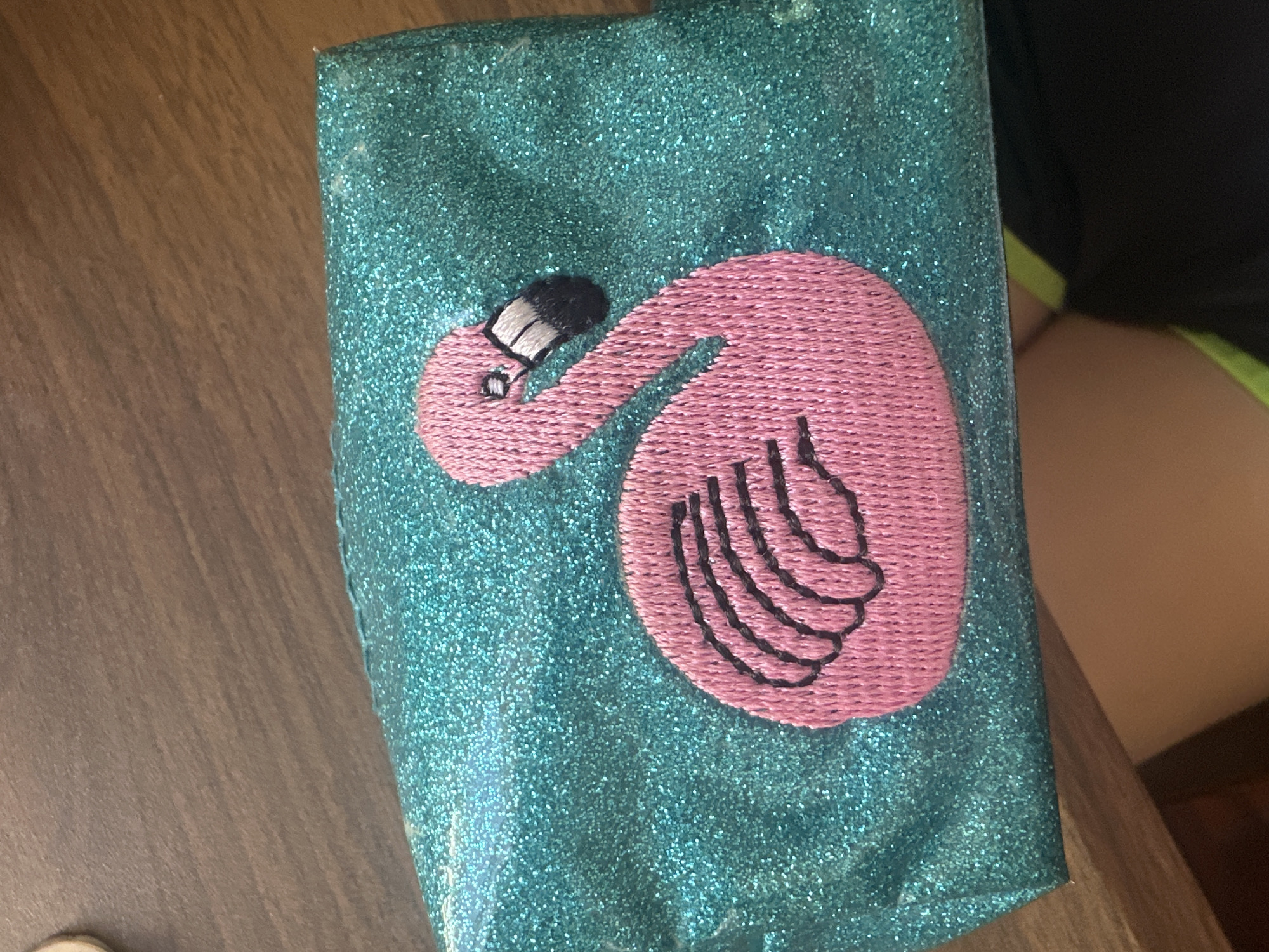 A close-up shot shows a teal, glittery pouch or case lying on a wooden surface. Embroidered on the pouch is a pink flamingo with black details outlining its beak and feathers on its body. The overall composition is focused on the texture and design of the pouch. There is no indication of the location being Sofia, Bulgaria.