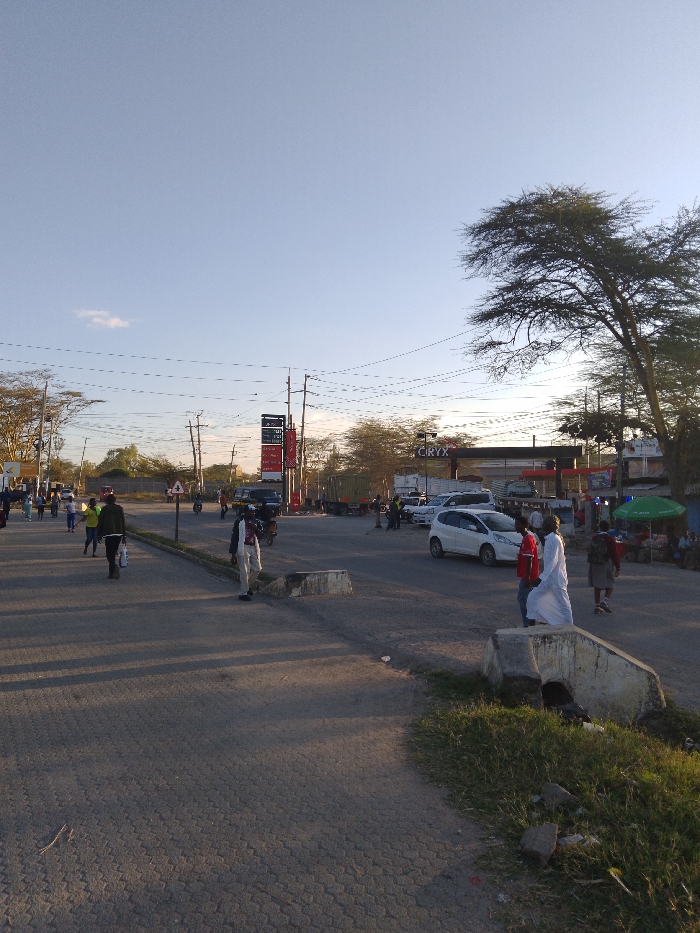 The image shows a street intersection in Athi River, Kenya, located at latitude -1.452 and longitude 36.98. The intersection is paved with asphalt and has a concrete curb on the right side. There are power lines overhead and a large tree on the right side of the image. In the distance, a gas station can be seen with a sign that reads "CIRYX". Several people are walking on the street, some wearing clothing, some wearing white robes. There are several cars parked on the side of the road, including a white hatchback. Some people are standing near the curb on the right side of the image, and some are walking in the road toward the camera. The sky is clear and blue with a few white clouds. The sun is shining, casting shadows on the ground. The environment is urban.