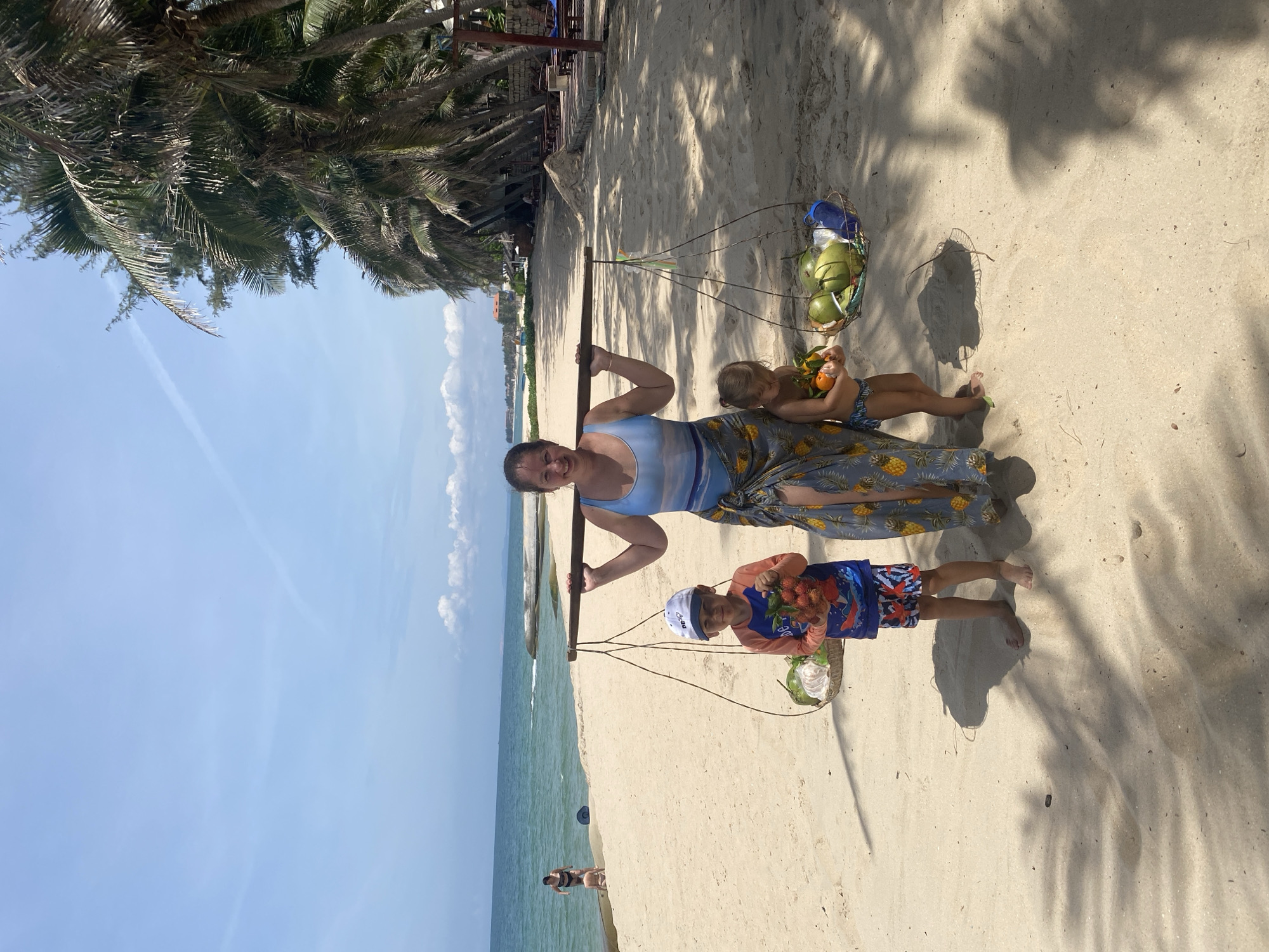 A woman, her son, and her daughter stand on a sandy beach, with palm trees lining the background. The woman is wearing a blue and white striped swimsuit and a patterned sarong. She is holding a shoulder yoke across her shoulders, with baskets of fruit hanging from each end. The son, wearing a blue rash guard and patterned swim trunks, stands to her left, holding a cluster of red fruits. The daughter, wearing a blue and white patterned swimsuit, stands to her right, holding an orange. The sea is visible in the background, with a few other people on the beach. The scene appears to be a hot, sunny day. The location is identified as Phu Thuy Ward, Vietnam.