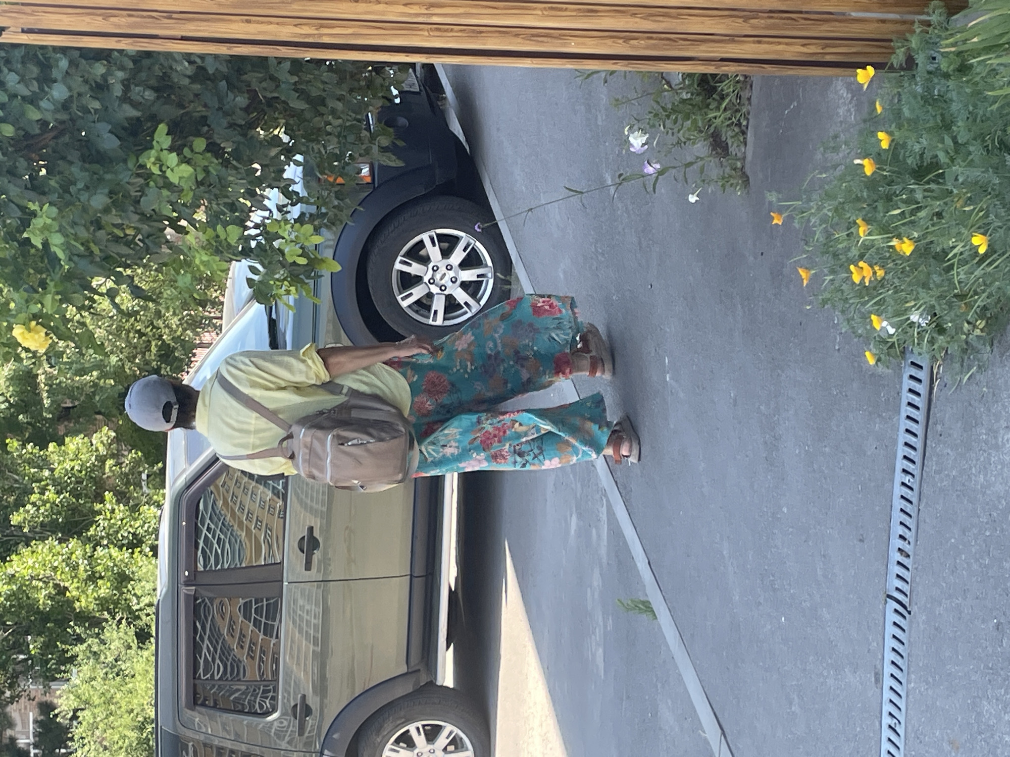 A person wearing a yellow shirt, floral pants, a grey cap, and sandals with a backpack walks on a paved area next to a dark green SUV. The SUV has tinted windows that reflect a building. In the foreground, there is a storm drain and a patch of yellow and white flowers. A wooden fence is visible on the right side of the image. The scene appears to be a street in Azov, Russia.