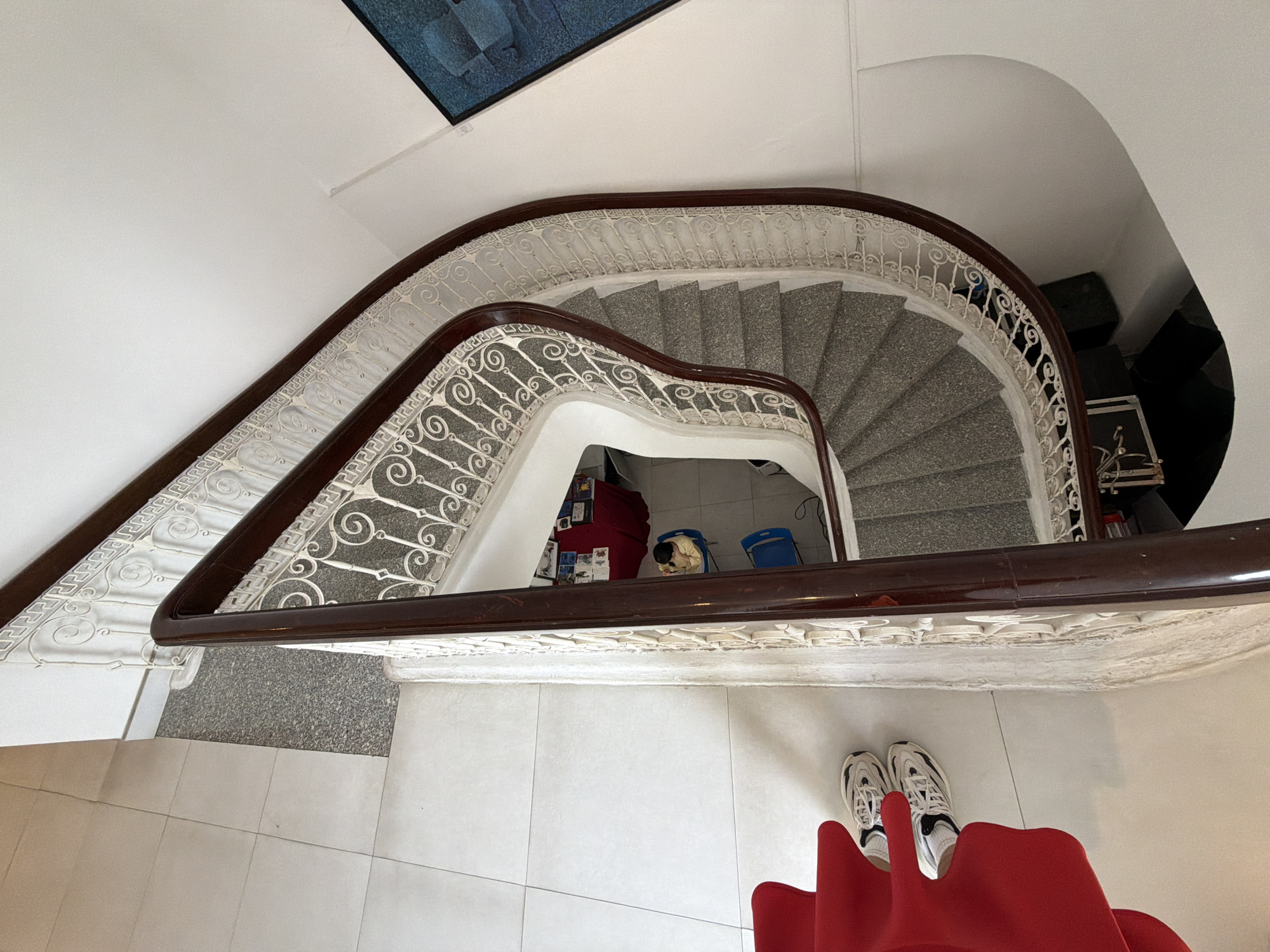A high-angle shot shows a curved staircase with ornate white railings and a dark brown handrail. The steps are made of grey speckled material. To the left of the staircase, a section of white tiled floor is visible, with a red chair partially in frame and a pair of white sneakers on the floor. In the background, near the landing of the staircase, a red table is set up with items on it, and two blue chairs are present, with a person wearing a yellow top sitting in one. Further down, a dark blue bag is visible. To the right of the staircase, a white wall displays a framed artwork with blue tones. The scene appears to be indoors, possibly in a public building or residence, and does not provide specific information about the city of Thuận Hóa Ward, Vietnam.