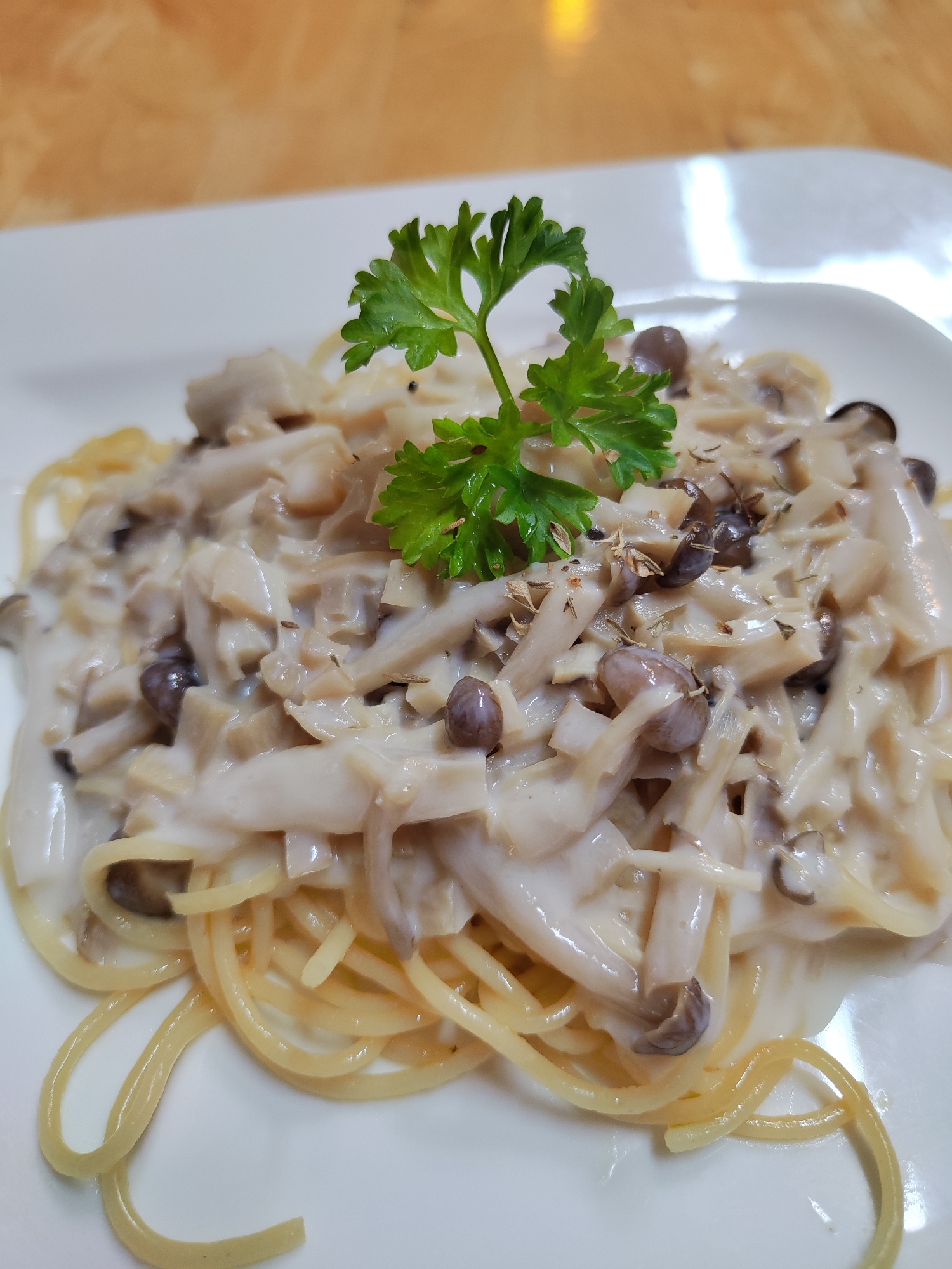 A close-up shot presents a plate of mushroom spaghetti, garnished with a sprig of parsley. The spaghetti is topped with a creamy mushroom sauce, featuring a mix of sliced and whole mushrooms. The dish is served on a square white plate. The background features a light brown, textured surface.