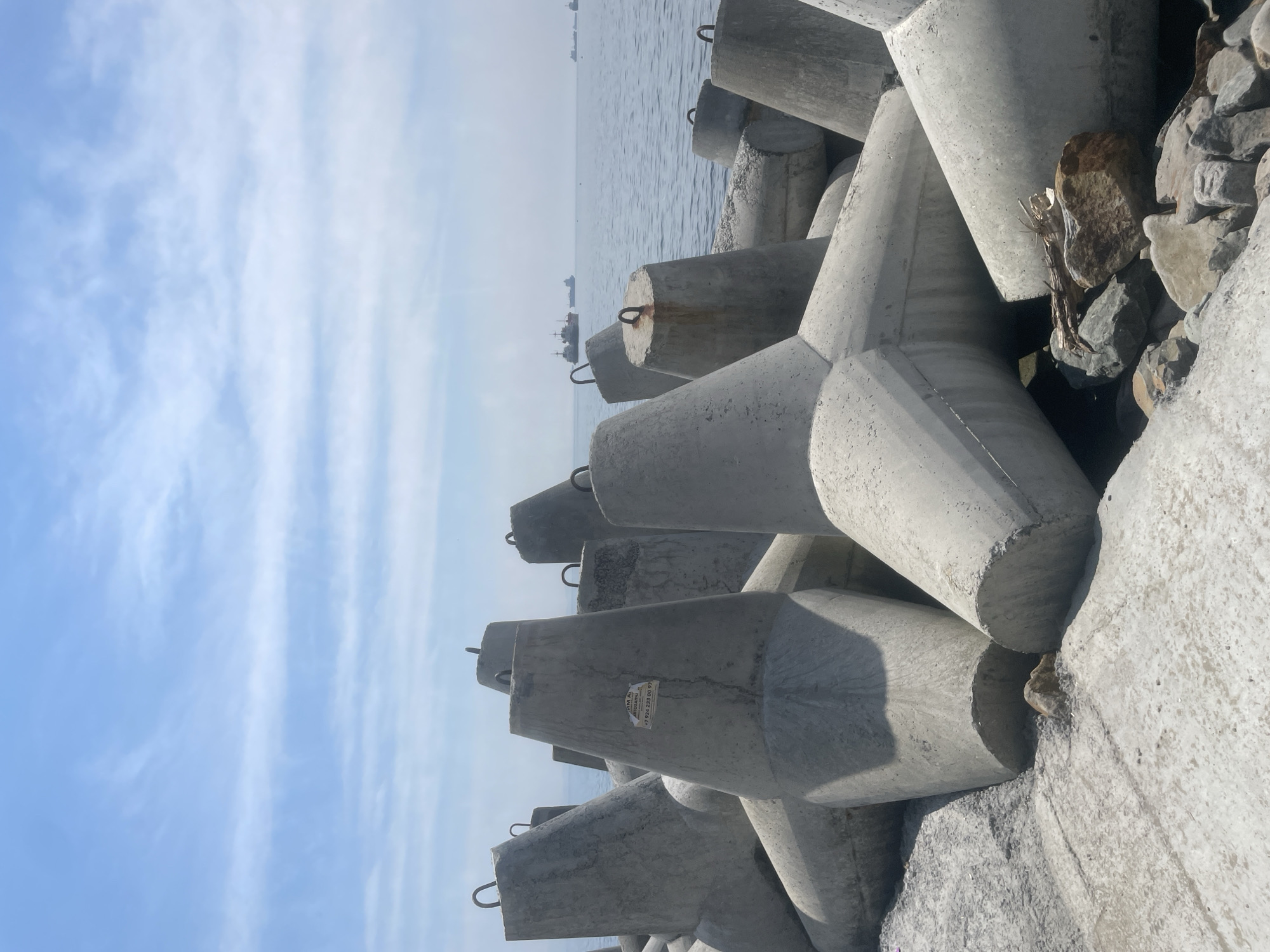 The image depicts a close-up view of concrete tetrapods arranged on a shore, likely serving as a breakwater. The tetrapods are massive, interlocking concrete structures designed to dissipate wave energy. They are gray and appear weathered, with visible seams and some rough edges. A few of the tetrapods have metal loops protruding from their tops, used for lifting and placement. One tetrapod in the foreground has a sticker on its side with Cyrillic text and a phone number, suggesting a commercial or construction context.
Beyond the tetrapods, the sea stretches out to the horizon under a bright, partly cloudy blue sky. Several cargo ships and other vessels are visible on the water, indicating an active shipping lane or port area. The shoreline itself is a mixture of concrete surfaces and rough rocks. In the lower right corner, a piece of driftwood rests among the rocks, adding a natural element to the industrial scene. The overall impression is one of coastal engineering at a busy maritime location.
This scene is characteristic of the breakwaters found along the coastline of Vladivostok, Russia, a major Pacific port city. The presence of large ships and the type of coastal defense structures strongly suggest a location within or near the port facilities of Vladivostok.
