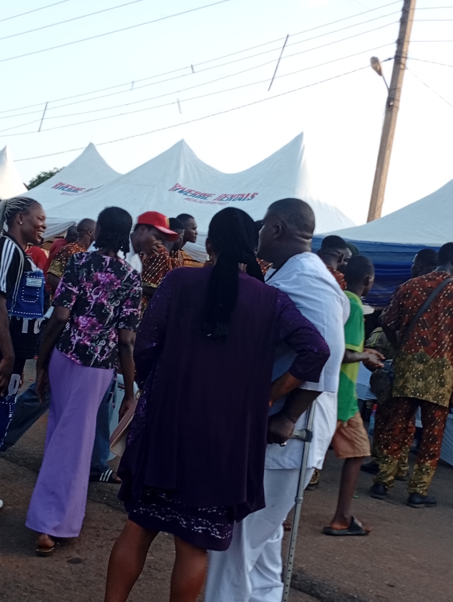 The image depicts an outdoor gathering near tents, possibly for an event, with a large number of people. In the foreground, a woman in a purple dress is seen from behind, facing away from the camera. To her left, another woman in a black and white striped shirt and a blue vest is laughing. A man in a white outfit and a woman in a purple skirt are also visible in the foreground. In the background, there are more individuals, some wearing traditional African attire. Tents with "RIVERINE RENTALS" written on them are set up behind the crowd. A utility pole with a light fixture is visible on the right side of the image. The ground appears to be asphalt. The setting suggests a community event or celebration in Nsugbe, Nigeria.
