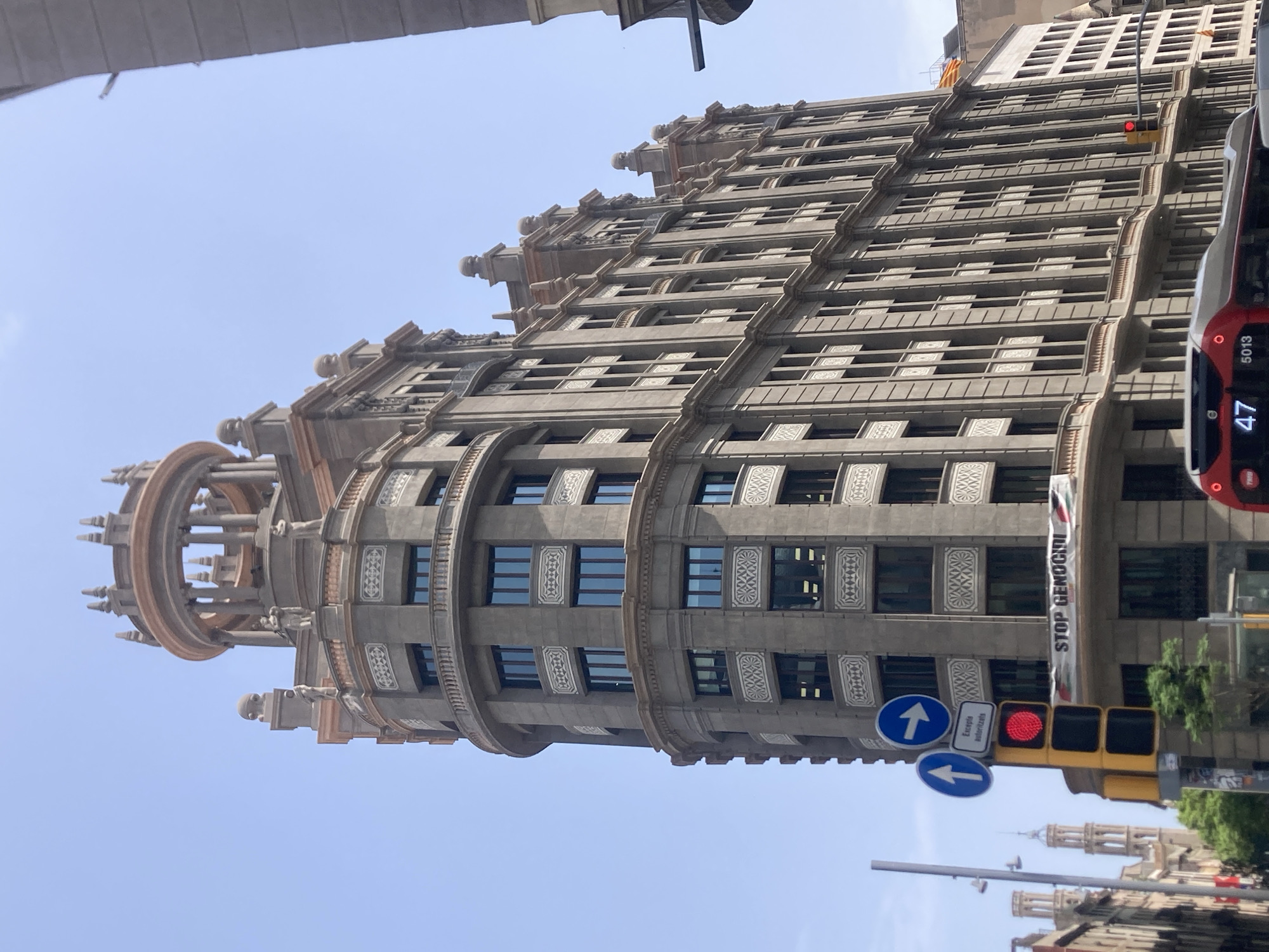 The image captures a wide-angle view of a grand, multi-story building in Barcelona, Spain, under a clear blue sky. The building, with its ornate architectural details, features a prominent rounded facade on the left and a more linear section on the right. Decorative elements such as carved reliefs, balconies with railings, and a domed cupola at the very top are visible.
In the foreground, a traffic light displays a red signal. Below the traffic light, a banner is hung across the building, displaying the text "STOP GENOCIDE" alongside images of what appears to be a flag. Several road signs are also present, including one with an upward-pointing arrow and another with a rightward-pointing arrow, along with a smaller sign that reads "Excepte autoritzats."
A red bus with the number "47" and "5013" displayed on its front is visible at the bottom of the frame, suggesting it is stopped at the traffic light. In the background, the spires of other buildings can be seen, adding to the urban landscape of Barcelona. The perspective suggests the photo was taken from street level.