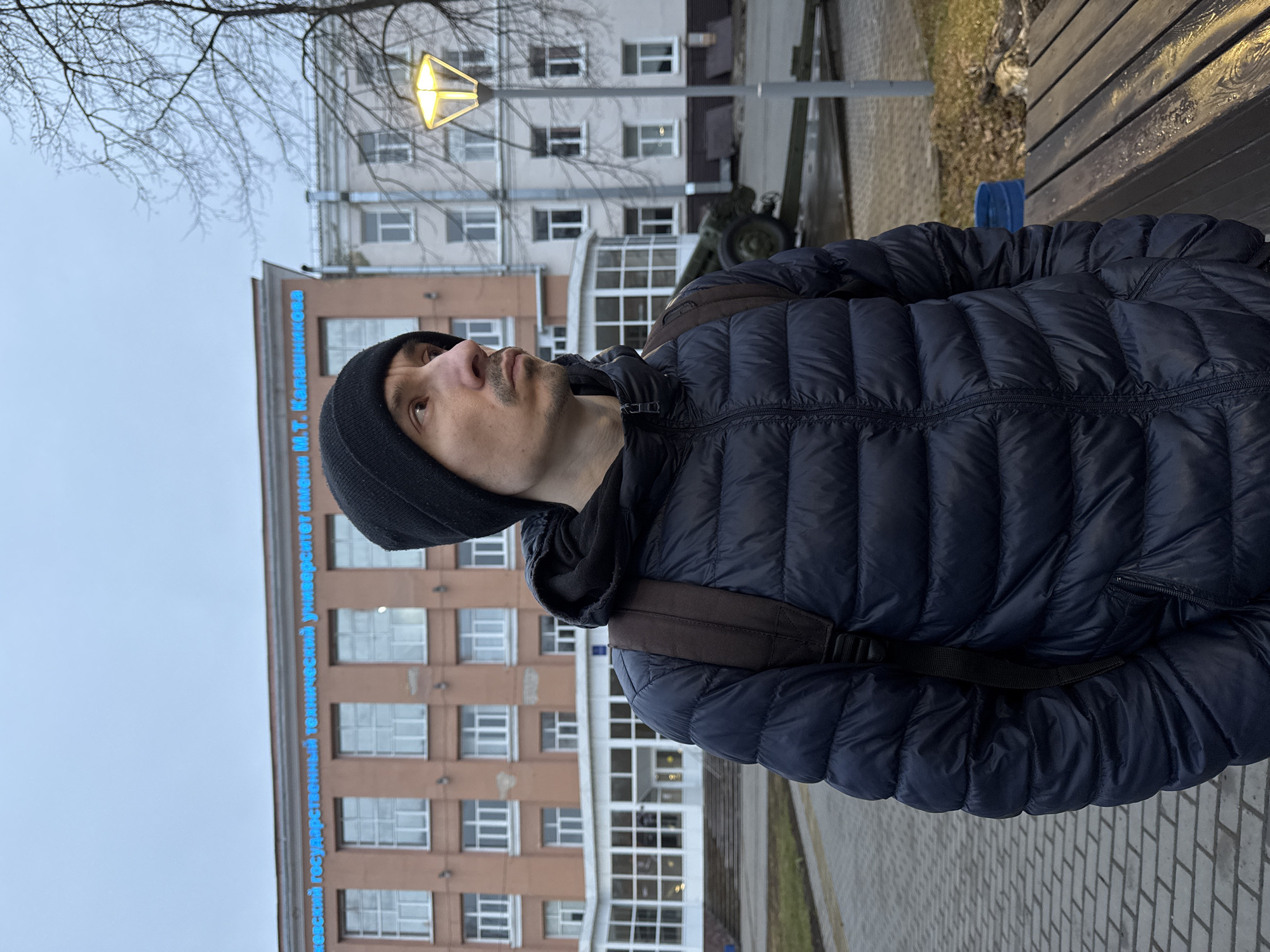 A man with a black beanie and a dark blue puffy jacket with a backpack stands on a paved area. He is looking upwards. Behind him is a large building with numerous windows. Blue writing is visible along the building's facade, identifying it as Izhevsk State Technical University named after M.T. Kalashnikov in Izhevsk, Russia. In the background, there is a street lamp and a cannon on display. Part of a wooden bench is visible on the right. The sky is overcast.