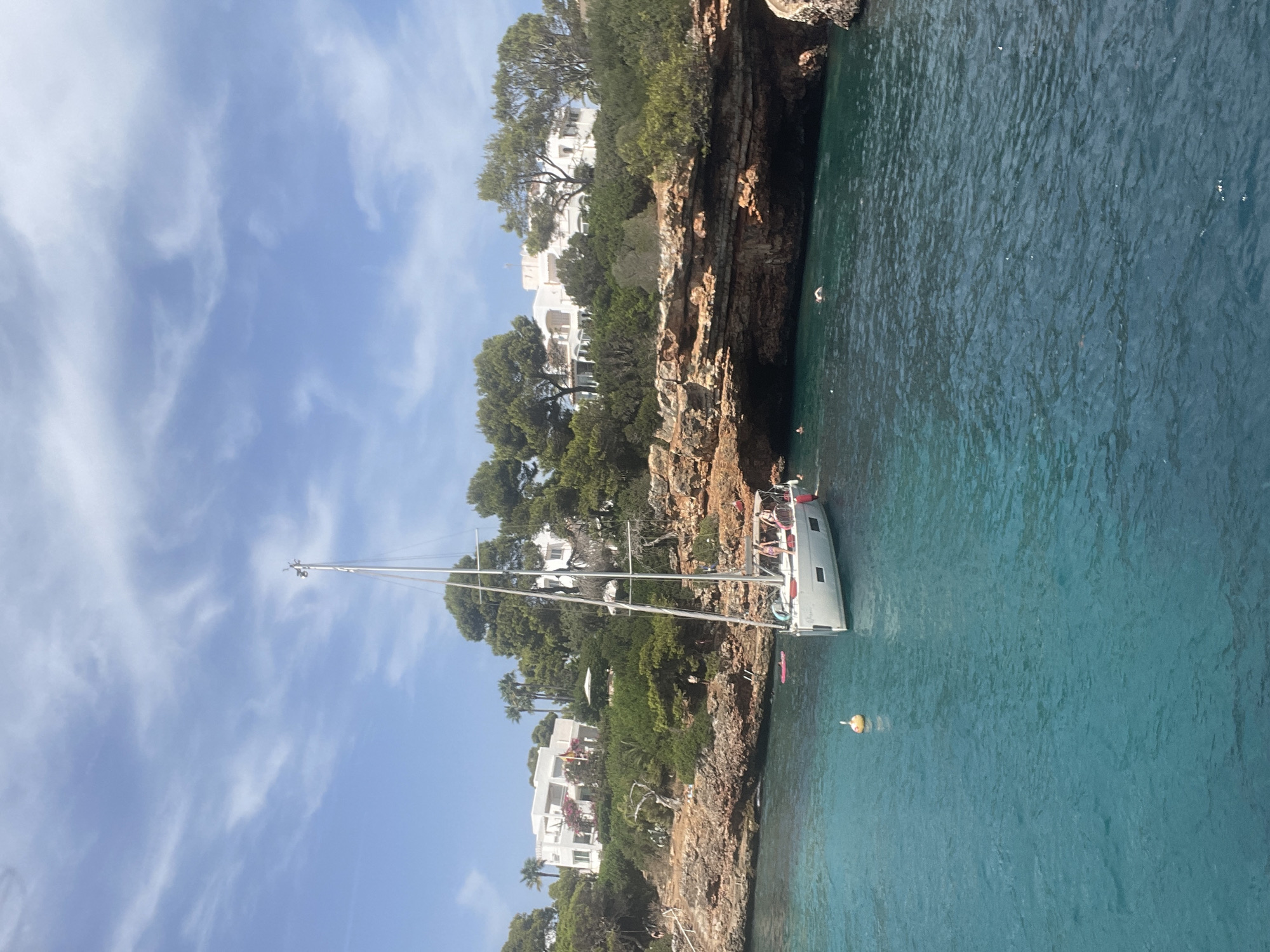 The image depicts a scenic coastal view in Santanyí, Spain, specifically near Cala Llombards. A white sailboat is anchored in the clear, turquoise waters of the Mediterranean Sea, close to a rocky coastline. Several people are visible, some swimming in the sea and others on the shore or on the boat. The coastline is characterized by rugged, brown rocks covered with lush green vegetation and trees. In the background, white buildings with red-tiled roofs are nestled amidst the trees, suggesting a residential or resort area. The sky is blue with scattered white clouds. A yellow buoy floats in the water near the sailboat.