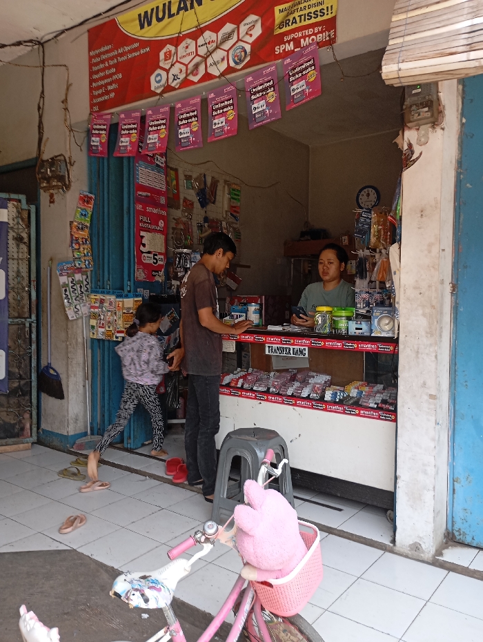 The image shows a small shop in Katapang, Indonesia (latitude: -6.99, longitude: 107.552). The shop is brightly lit and has a large sign above the entrance advertising "WULAN" and "Unlimited" mobile phone packages. The shop is filled with various products and merchandise, including phone accessories and prepaid cards.
Two people are present in the shop: a man in a blue shirt and jeans, standing near the counter and looking at something in a shelf, and a woman in a blue shirt and black skirt, standing behind the counter and looking at something on her phone.
A small child, wearing a pink shirt and blue shorts, is standing in the left side of the image, looking at something in a display rack behind the counter. The child is wearing a backpack and holding a red bag in their right hand.
The image is taken from the left side of the shop. The floor is tiled and clean. A small pink and white bicycle is partially visible in the bottom right corner.