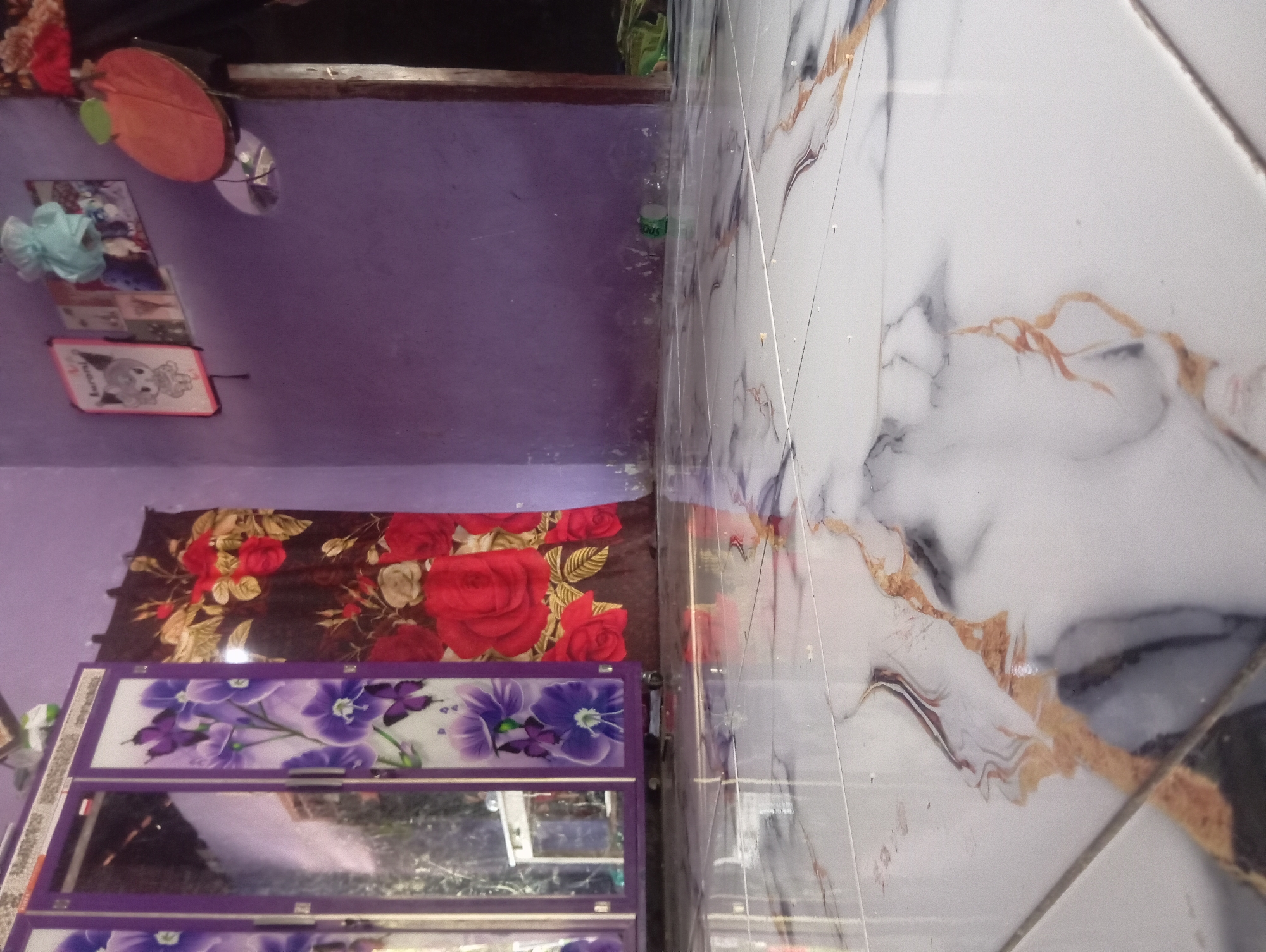 The image shows the interior of a room with purple walls and a marble-patterned tiled floor. On the left, there is a purple wardrobe with glass doors featuring floral and butterfly designs. To the right of the wardrobe, a brown curtain with red floral patterns is visible. On the purple wall, several framed pictures and a circular orange decoration are hung. The foreground is dominated by the reflective tiled floor. The scene is likely within a residential setting in Dente Teladas, Indonesia, based on the provided context. There are no people visible in the image.