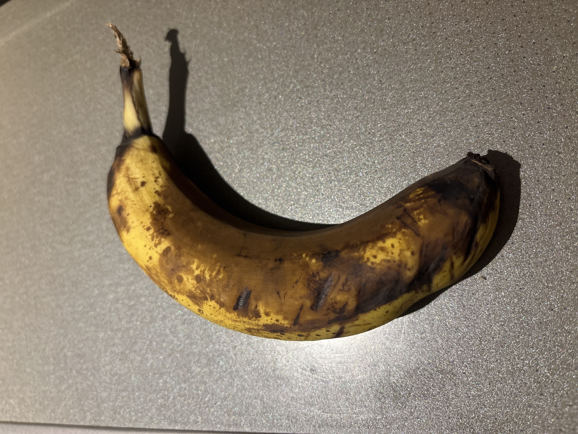 A slightly overripe banana with brown spots and some dark bruises lies on a speckled gray countertop. The banana is positioned horizontally, curving slightly upwards from left to right. The stem end is to the right and appears somewhat dry and frayed. The lighting casts a shadow to the lower right of the banana. The environment appears to be an indoor setting, likely a kitchen. There is no indication of any specific location in Dubai, United Arab Emirates.