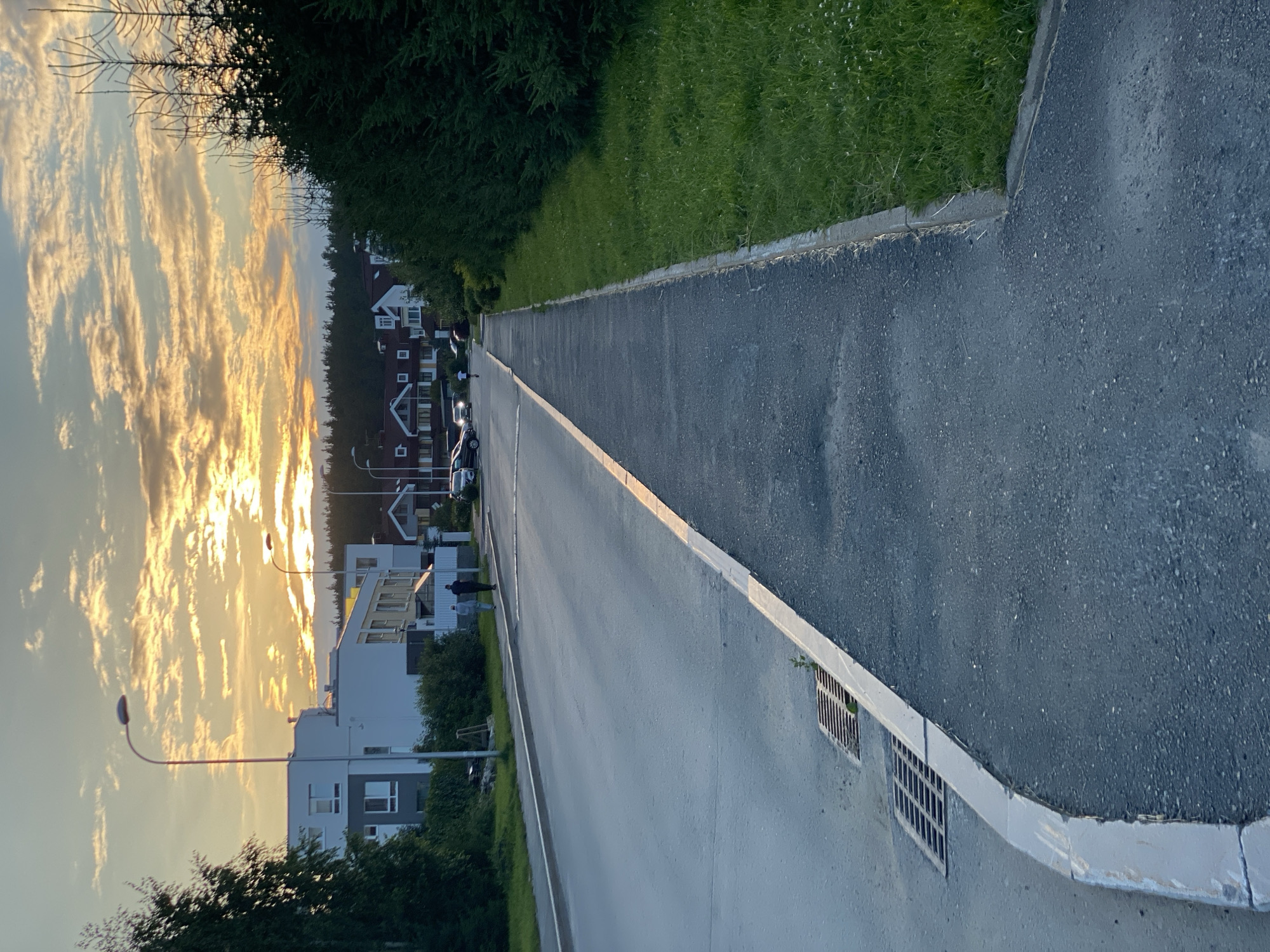 This is a vertical, eye-level, outdoor, daytime photograph of a street in a residential area, likely in Yekaterinburg, Russia. The sun is setting, casting a warm, golden glow on the sky, which is filled with dramatic, wispy clouds. The light illuminates the tops of the trees and the roofs of the houses on the left side of the street.
The street itself is paved with asphalt and appears to be a residential road. There is a white line marking the edge of the road, and a curb separating it from a grassy area on the left. Several modern-looking houses with red and white facades are visible, suggesting a contemporary housing development. Some of these houses have multiple stories and balconies.
Streetlights with curved poles are present along the street, one of which is illuminated. There are a few cars parked along the road. In the middle distance, two figures are walking on the sidewalk, and another is standing near a house. A drainage grate is visible along the edge of the road on the right.
The overall mood of the image is peaceful and serene, enhanced by the warm sunset light and the quiet atmosphere of the residential street. The weather appears to be clear and calm. The scene depicts a typical evening in a suburban or newly developed urban area.