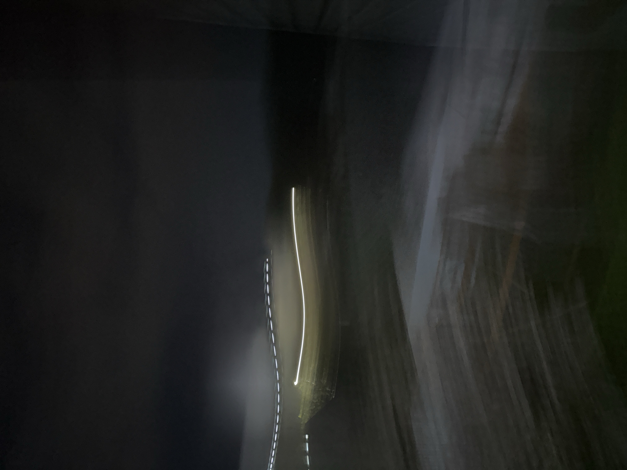 The image shows a dark, blurry scene with light trails. A distinct white light trail curves vertically in the middle. Dashed white lines are visible to the left of the solid light trail. The left side of the image is darker and appears to be a solid surface. The right side of the image shows blurred vertical streaks of grey and green, suggesting motion or a long exposure.
It is not possible to determine the specific location in Nizhny Novgorod, Russia, without additional context or information. No people, subjects, objects, actions, or interactions are evident in the image beyond the described light trails and blurred environment.