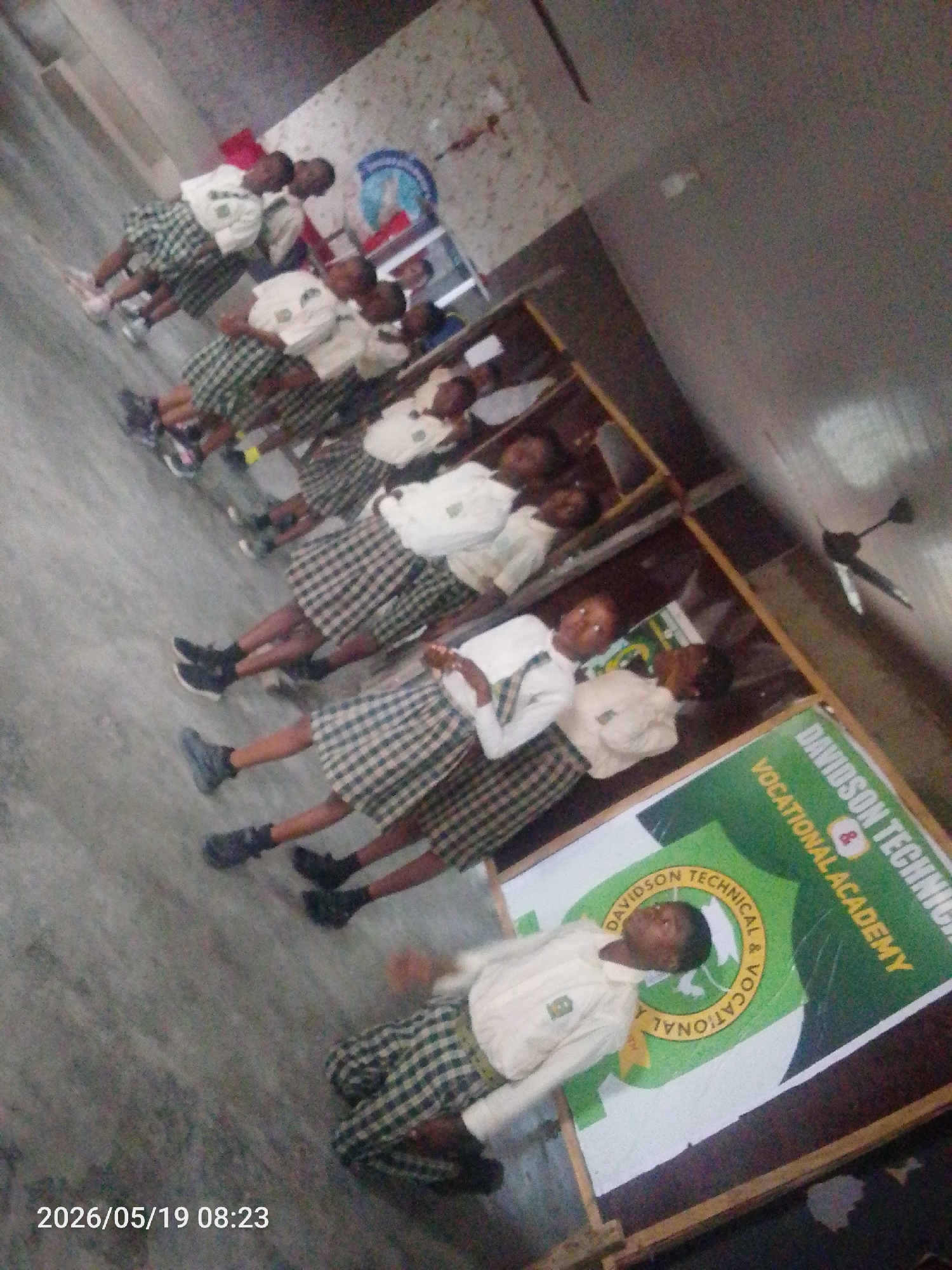 The image, dated 2026/05/19 08:23, depicts a group of young people, likely students, standing in a line. They are wearing what appears to be a school uniform consisting of white shirts and plaid skirts or shorts. Behind them is a sign that reads "DAVIDSON TECHNICAL & VOCATIONAL ACADEMY." The setting appears to be indoors, potentially a school hallway or classroom in Lokoja, Nigeria. The individuals are mostly looking towards the camera. The image is somewhat grainy and taken at an angle. A ceiling fan is visible in the upper right corner. The flooring seems to be gray or concrete.