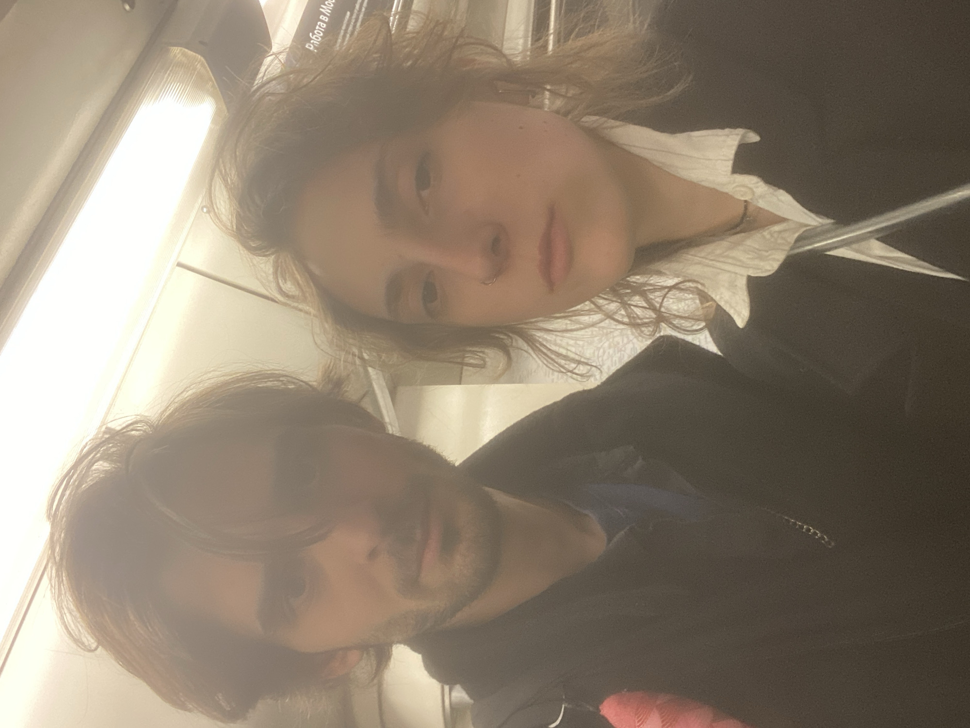 Two people are on the Moscow Metro. The first person is on the top of the image and is fair-skinned with light brown hair. They are wearing a white shirt with a dark jacket. The second person is at the bottom of the image and is a fair-skinned male with brown hair and a beard. He is wearing a black jacket and a blue shirt. They are inside a subway car with white walls and a light fixture.