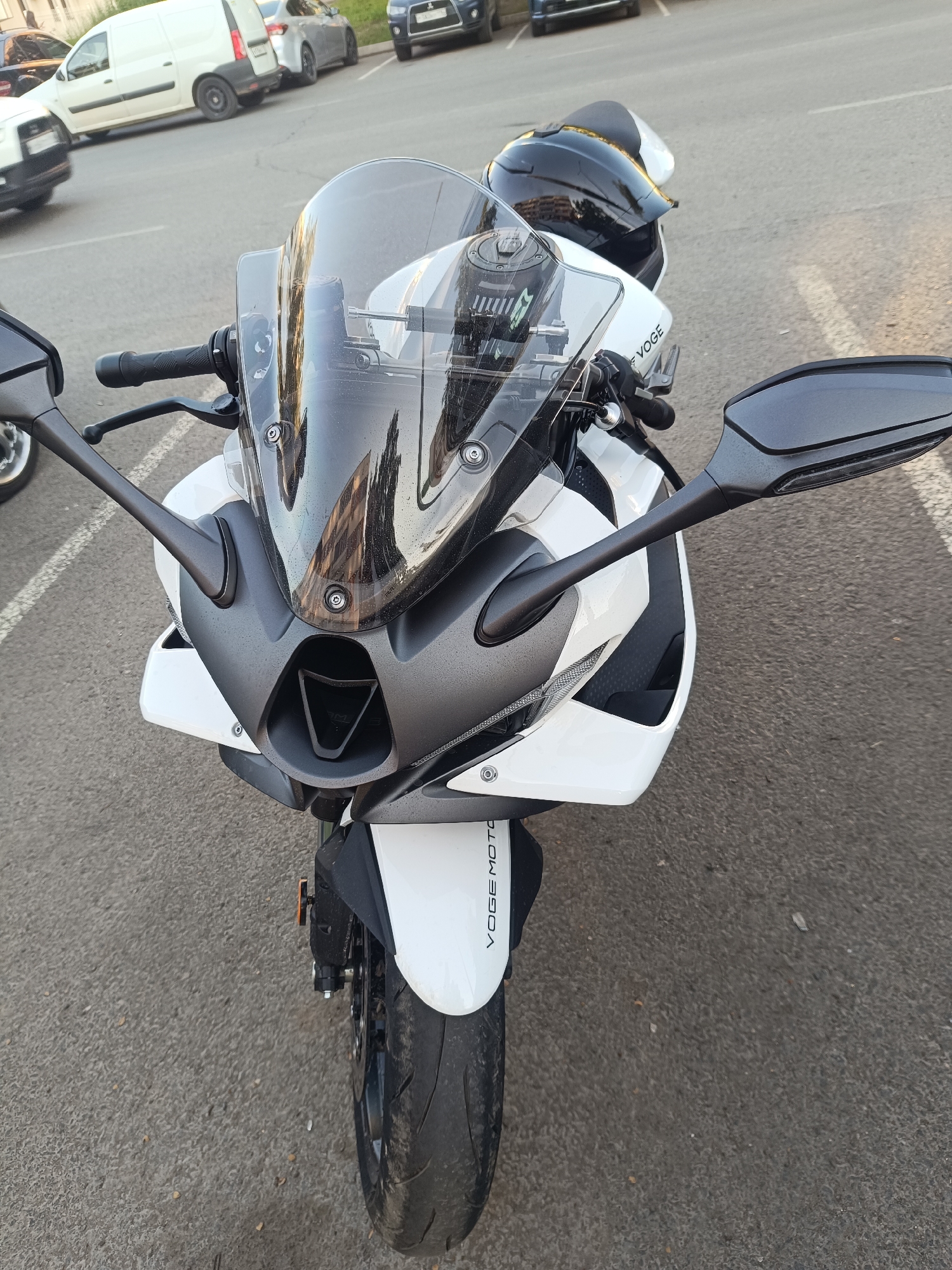 The image shows the front of a white and grey sport motorcycle parked on an asphalt surface, possibly a street or parking lot, in Naberezhnye Chelny, Russia. The motorcycle has a dark tinted windshield, black handlebars, and mirrors. A black helmet is visible on the seat of the motorcycle. In the background, several parked cars and a white van are visible, along with markings on the asphalt that suggest a parking area. The lighting suggests it is daytime, likely late afternoon or early evening due to the warm tones in the reflection on the windshield. There are no people visible in the image. The overall scene depicts a parked motorcycle in an urban environment.