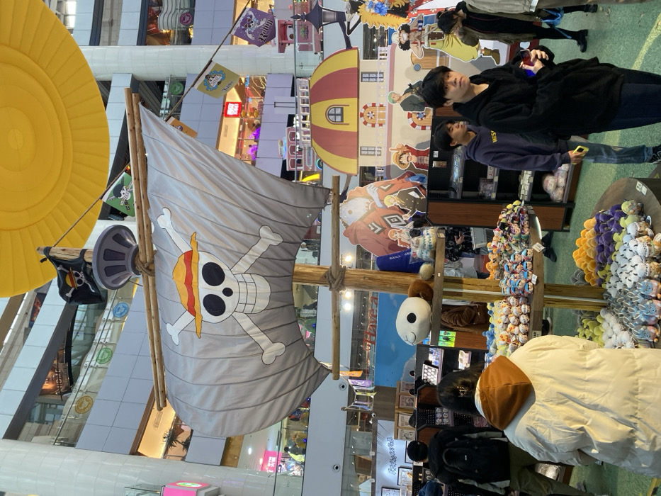 This media file captures an indoor commercial space, likely a shopping mall or a themed exhibition area in Guang Zhou Shi, China, dedicated to the popular anime "One Piece." The image is presented in a vertical, portrait orientation.
In the mid-ground, a prominent, large-scale replica of a pirate ship's sail is featured, displaying the iconic Straw Hat Pirates Jolly Roger (a skull and crossbones wearing a straw hat). This sail is attached to a wooden mast structure. To its left, a smaller black flag also bears a pirate skull design. Above the sail and to the left, a large, bright yellow, circular inflatable object is visible, possibly part of a hot air balloon or another decorative element.
Several people are present, indicating a public event or shopping activity. In the mid-ground, two young men with dark hair are looking towards the camera. The man on the right wears a black hoodie, and the one behind him wears a dark blue hoodie. In the foreground, a person in a cream or white puffer jacket is seen from behind, looking at merchandise. Other blurry figures can be discerned in the background.
The area is filled with One Piece-themed decorations and merchandise. Behind the people, cardboard standees of various One Piece characters, including Monkey D. Luffy, Roronoa Zoro, and Sanji, are set up. A multi-level structure with a distinctive red and yellow striped dome top resembles a building from the series. To the right, multiple merchandise stalls are laden with numerous plush toys of various colors like purple, yellow, white, and grey. A large white plush head, possibly a character, is also visible.
The activity appears to be people browsing and enjoying a themed retail or promotional event. The setting suggests a vibrant atmosphere with bright indoor lighting. Visible text includes "Rulun" and "Totor" on small flags, and "SANJI" on a character cutout. The overall scene highlights a lively fandom event.