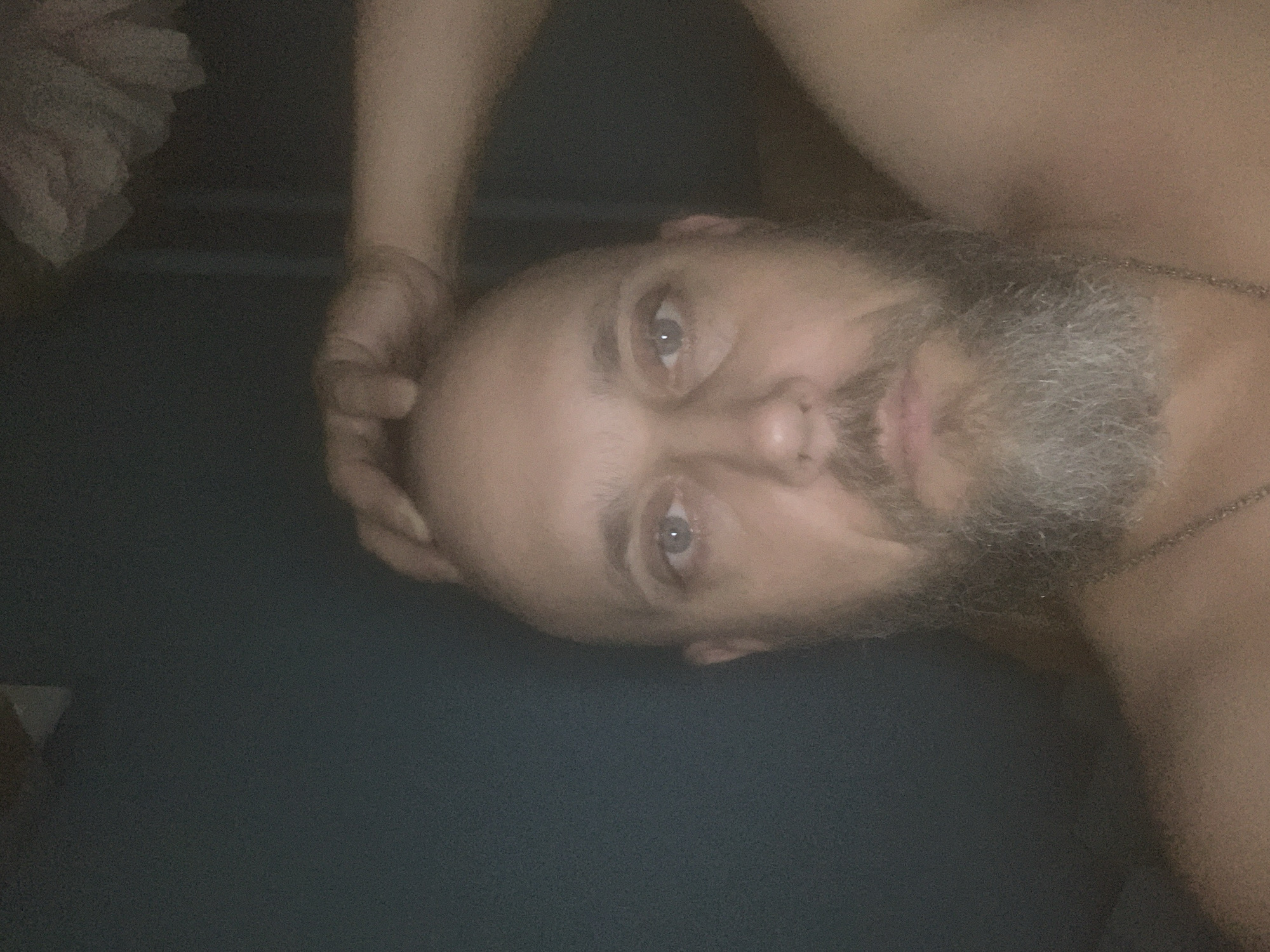 A close-up, over-the-shoulder selfie taken in a dimly lit indoor setting. The subject is a bald man with a grey beard, positioned in the center of the frame. His left eye is visible, looking towards the camera. His left hand is raised and touching the top of his head. The background is a dark, solid color, possibly a wall or curtain. There is a hint of a white, textured object in the upper right corner. The man is wearing a necklace. The image has a grainy quality due to low light conditions. There is no discernible indication of the specific location within Voronezh, Russia.