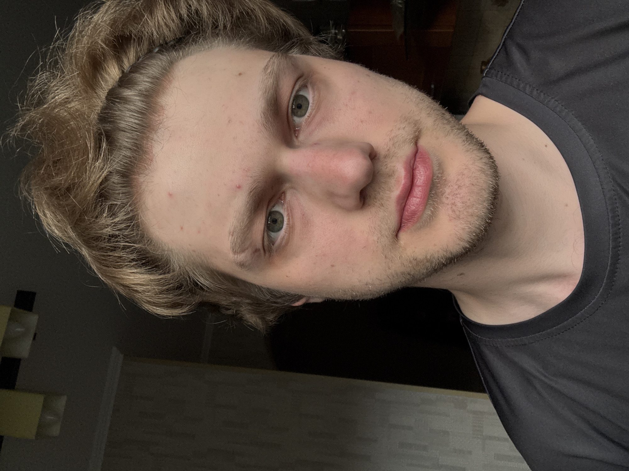 A close-up, waist-up selfie of a young Caucasian man with blonde, messy hair is taken indoors, likely in an apartment in Khimki, Russia. He is wearing a black t-shirt. His expression is neutral, with his gaze directed towards the camera. The background is dimly lit, with a textured wall visible on the left and a dark area on the right, possibly indicating a doorway or another room. Lighting appears to be from an overhead source. The overall mood is casual and candid.