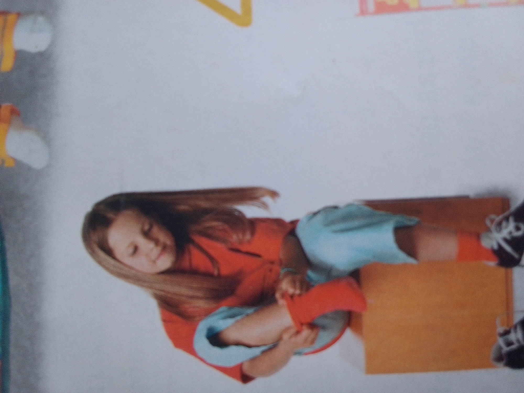 This is a vertically oriented, close-up, slightly blurry photograph featuring a young girl with long brown hair. She is wearing a red shirt and light blue shorts. The girl is sitting on a wooden surface, possibly a bench or a low table, which is a warm, orange-brown color. Her left leg is bent, and she appears to be adjusting a red sock or shoe on her right foot. Her expression is relaxed, with her eyes closed or looking down. To the right of the girl, partially visible, are a pair of black and white sneakers.
In the background, there are abstract elements that suggest a child-friendly environment. Yellow geometric shapes are visible at the top left, and there are also some blurry white and yellow objects that could be part of a toy or decoration. The overall background is a plain, light gray or white. The lighting is bright, suggesting it could be daytime. There are no obvious signs of weather or specific location cues beyond the implied indoor or set environment. The image conveys a sense of calm and everyday activity.