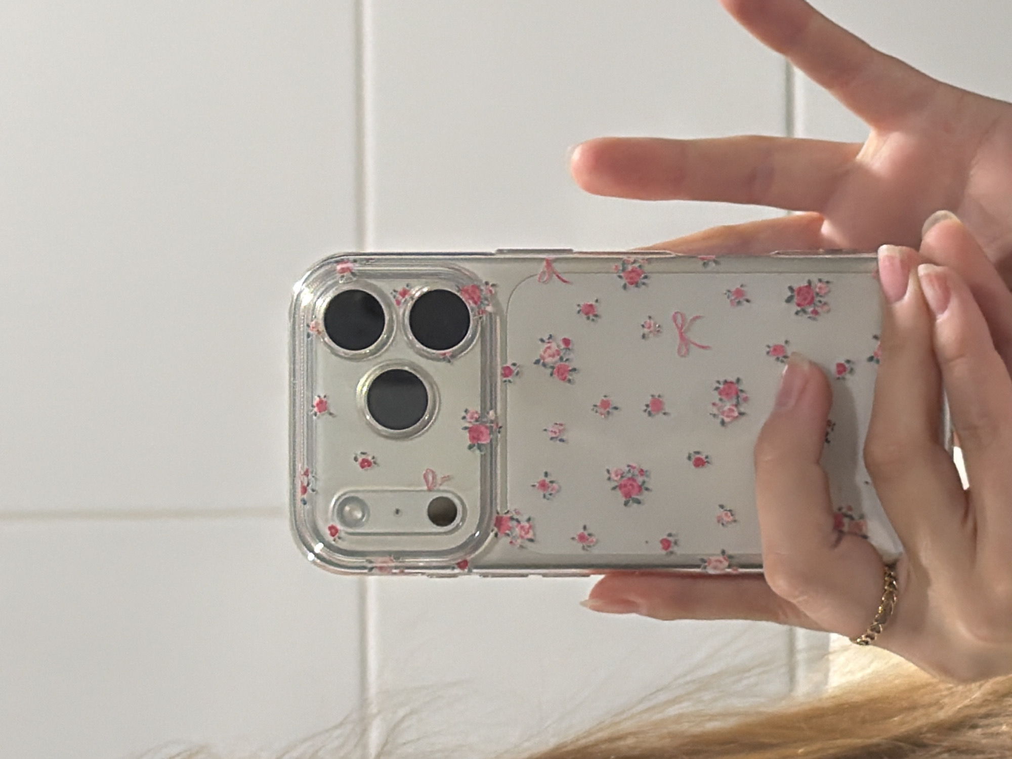 A person is holding a smartphone in their hands. The phone has a transparent case with a floral pattern. The case has a clear border and features pink roses and a pink ribbon design on a light background. The phone's camera module, with three lenses and a flash, is visible through the case. The person has a gold chain ring on their finger. The background is a white tiled wall. The location is not possible to determine based on the image.