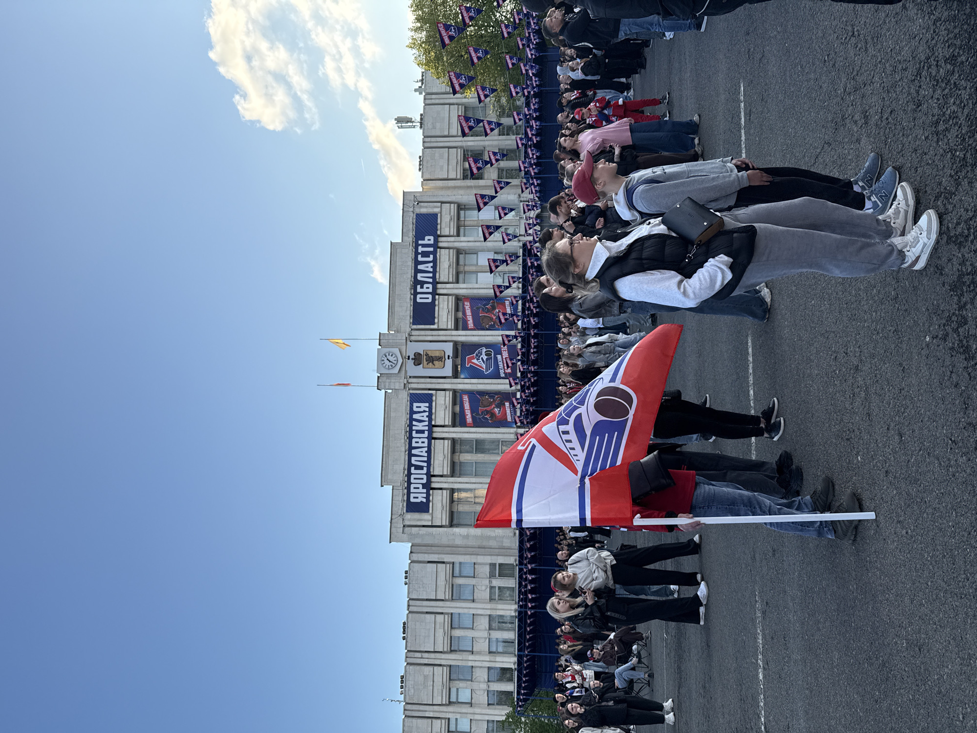 The image shows a crowd of people gathered in front of a large building with banners that read "ЯРОСЛАВСКАЯ ОБЛАСТЬ". A flag with a hockey team logo is prominently displayed in the foreground. People are standing, sitting in chairs, or lying on the pavement. The sky is blue with some clouds. The location is Шебунино, Russia.
