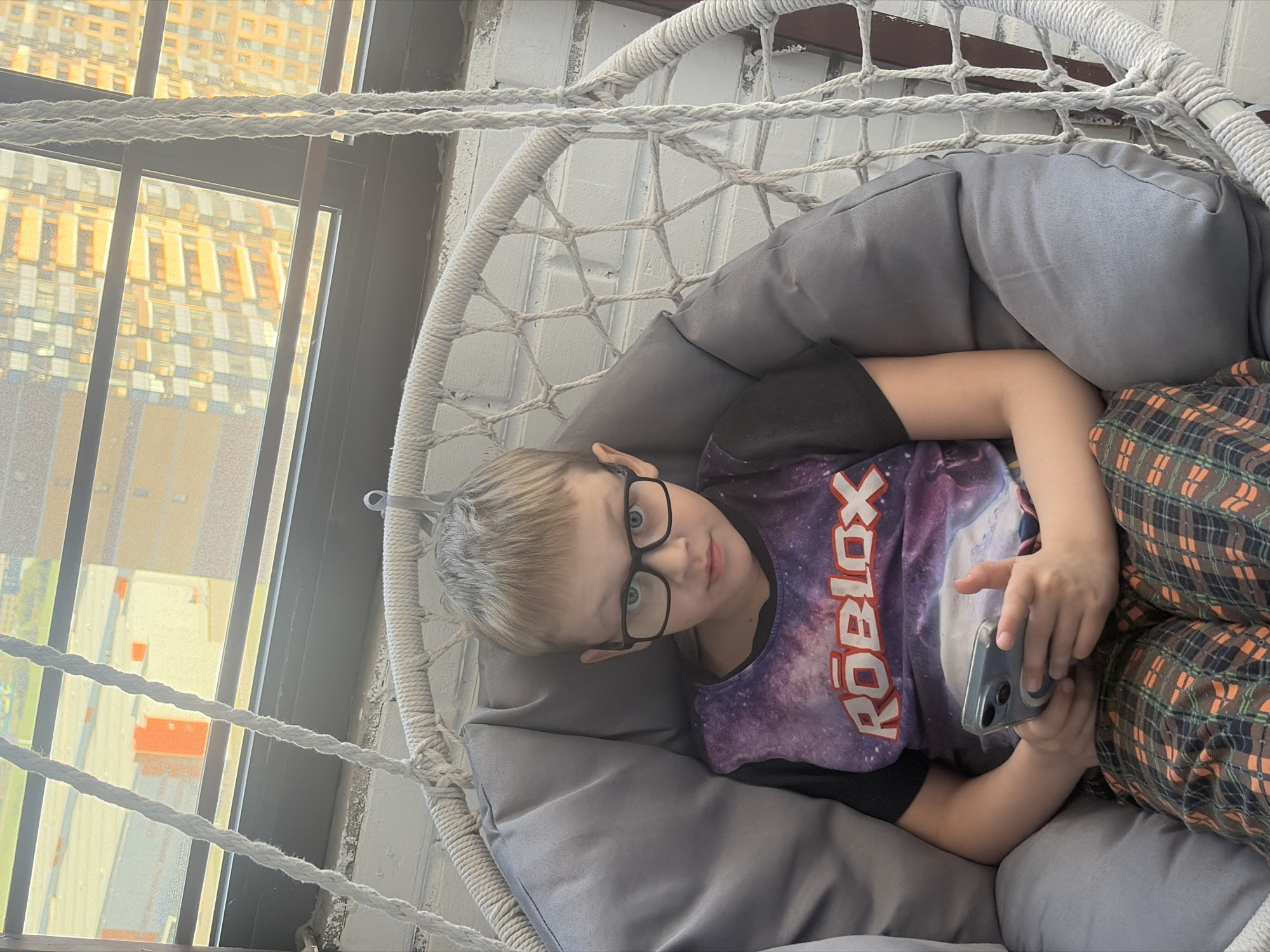 A young boy with blond hair and glasses is sitting in a gray cushioned swing chair. He is wearing a Roblox t-shirt and plaid pants, holding a mobile phone in his hands. The chair hangs near a window with a view of buildings, indicating a city location. The buildings are the same yellow and white color. Based on the building color and architecture seen through the window, the city is likely Saint Petersburg, Russia. The swing chair appears to be indoors, possibly in an apartment or home.