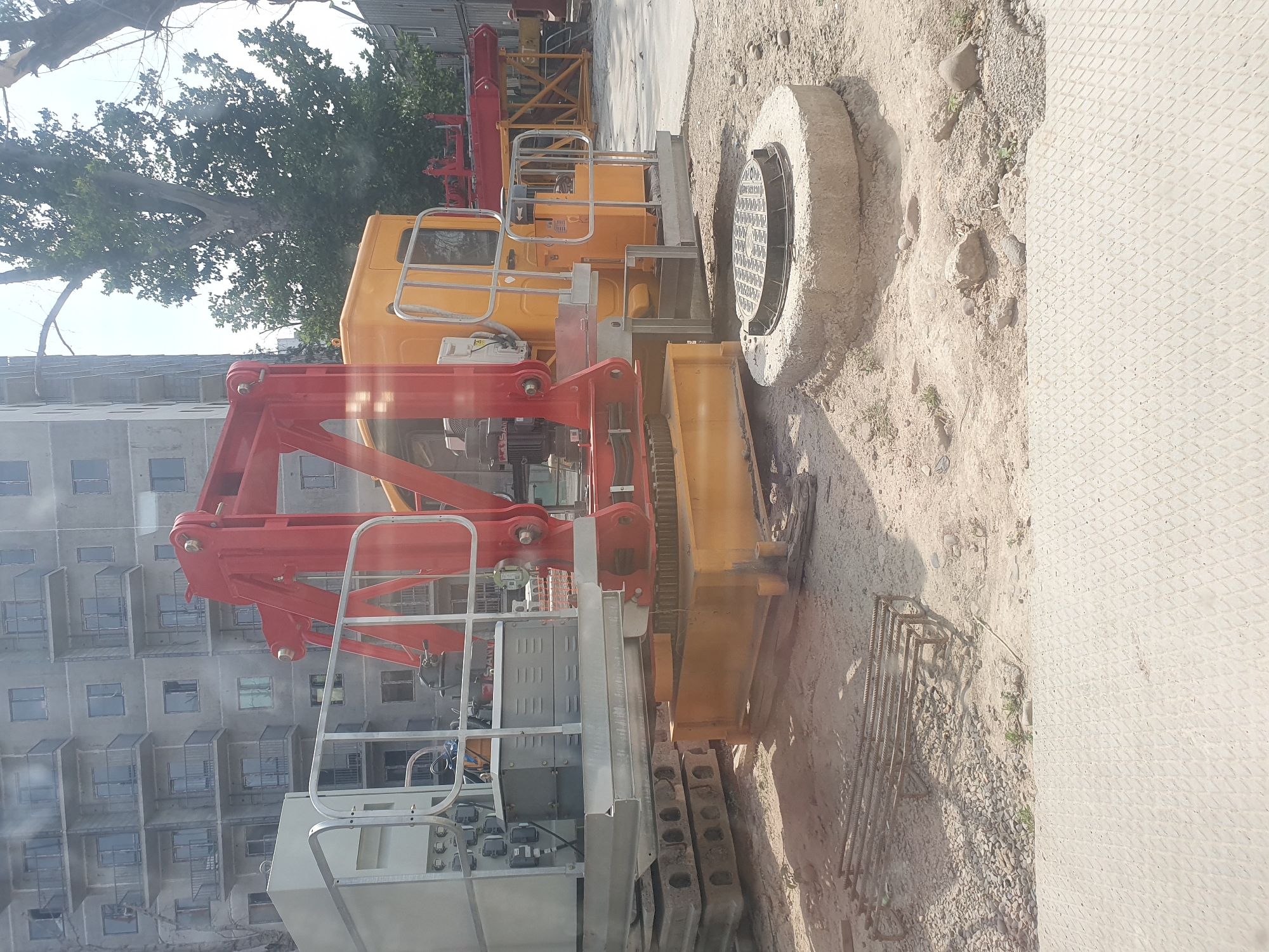 The image depicts a construction site in Tashkent, Uzbekistan. In the foreground, a large yellow and red crane mechanism is visible, resting on concrete slabs and a metal base. To the right, a manhole cover sits on a concrete ring surrounded by dirt and gravel. In the background, a multi-story apartment building with numerous balconies is under construction, and large trees with green foliage are present. The ground is a mixture of dirt, gravel, and some rebar reinforcements for concrete. The overall scene suggests ongoing construction activity in an urban environment.