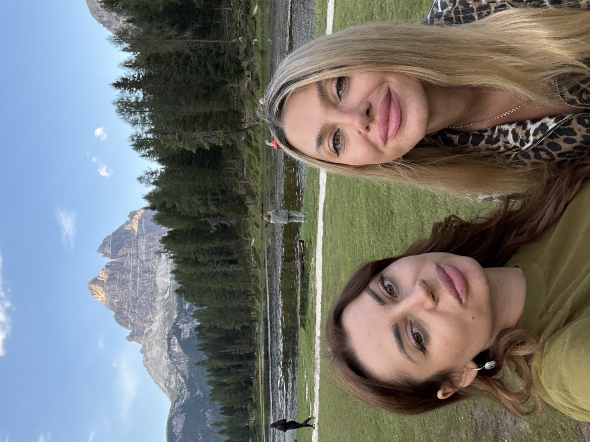 This is a selfie taken outdoors with two women in the foreground. The woman on the left has brown hair and is wearing an olive green top and a silver earring. The woman on the right has blonde hair and is wearing a leopard-print jacket. Behind them, there is a lake and a dense forest. In the background, a mountain range is visible under a blue sky with scattered clouds. The scene appears to be Lake Misurina, located near Auronzo di Cadore, Italy. There are a few other people visible in the distance near the lake and walking along a path. The overall atmosphere is serene and natural.