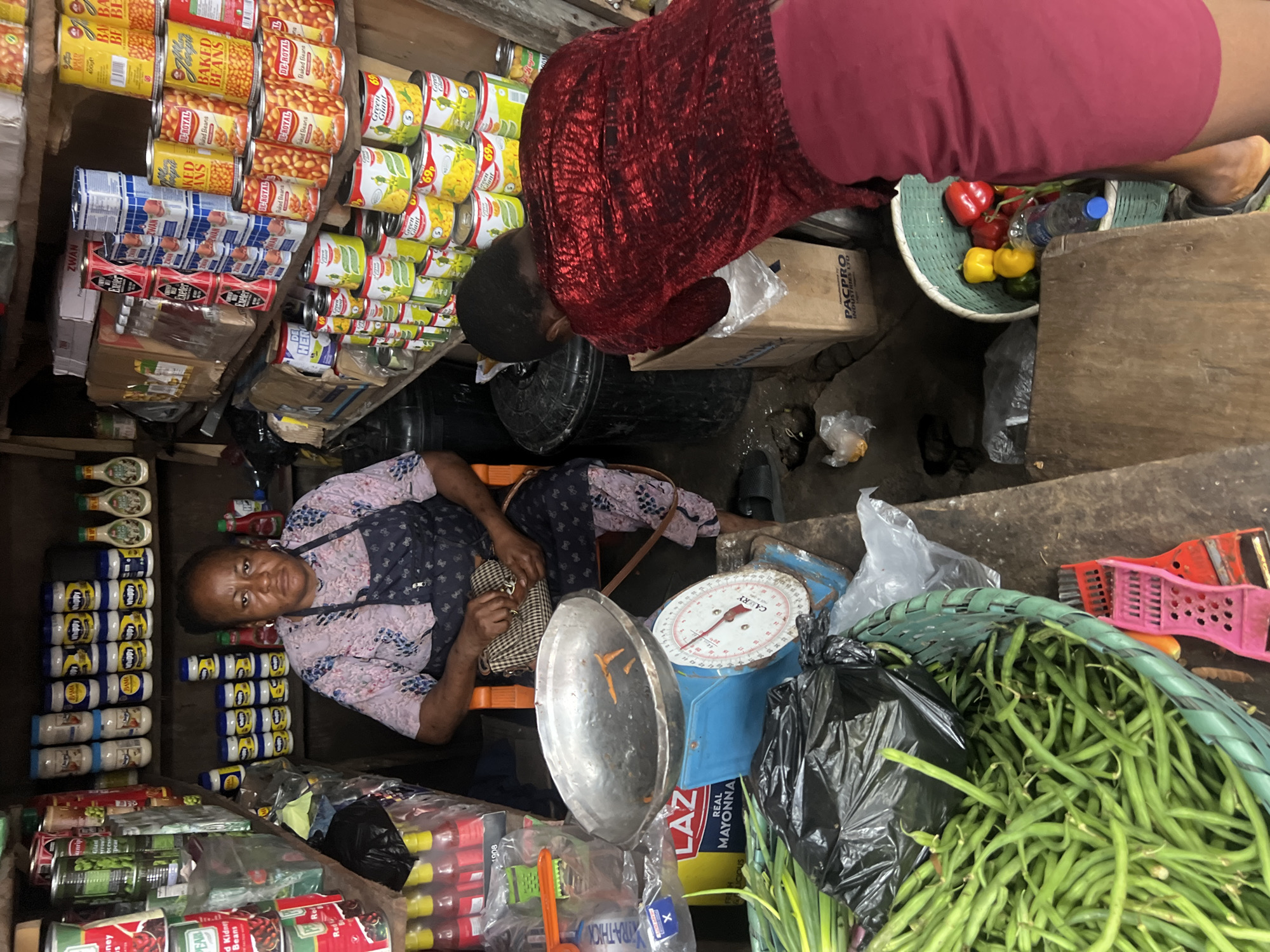 In Nkpor, Nigeria, a market scene unfolds with a woman at a stall. She is seated, wearing a light-colored floral top and a dark blue apron, with a checkered purse beside her. In front of her is a blue mechanical scale with a silver bowl. Beside the scale, a large green basket overflows with fresh green beans. To the right, a person wearing a sparkly red top and a maroon skirt is bending down, rummaging through a cardboard box. Behind the woman at the scale, shelves are packed with various canned goods, bottles, and jars. The stall appears cluttered, indicative of a busy marketplace environment.