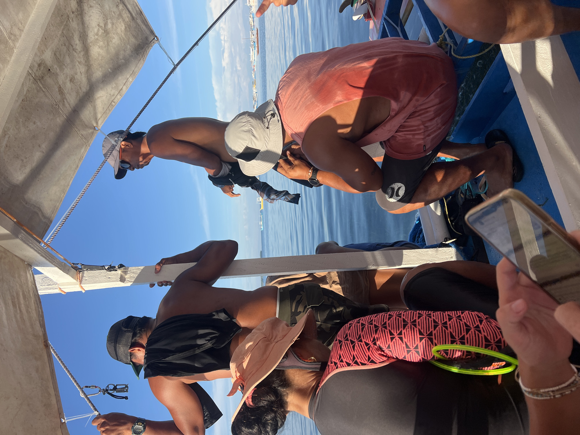 The image captures a scene aboard a traditional Philippine outrigger boat, likely in the waters off General Luna, Philippines, judging by the presence of multiple other boats and a hazy coastline in the background. The sky is a clear, bright blue, and the sea is calm and light blue.
In the foreground, partially obscuring the view, are the hands of a person holding a smartphone, with a silver bracelet on their wrist. The person is wearing a dark long-sleeved shirt with a distinctive pink and black geometric pattern on the sleeve, and a bright green ring or hoop is visible on their arm.
Seated on the boat's blue bench is a man wearing a light red sleeveless shirt and black shorts, with a bucket hat on his head. He is facing away from the camera and appears to be looking out at the water. Another man, also facing away, is standing and leaning against a white wooden post. He is wearing a baseball cap and has a black garment draped over his shoulder. His watch is visible on his left wrist. To his left, a woman in a pink sun hat and a dark-colored top with a similar geometric pattern to the person in the foreground, is also seated on the boat.
The boat itself is made of wood with blue and white painted surfaces. Ropes and a white canvas canopy are part of the boat's structure. Several other boats, including some with sails, are visible in the distance on the water. The overall atmosphere is one of a sunny day at sea, possibly during a boat trip or excursion.