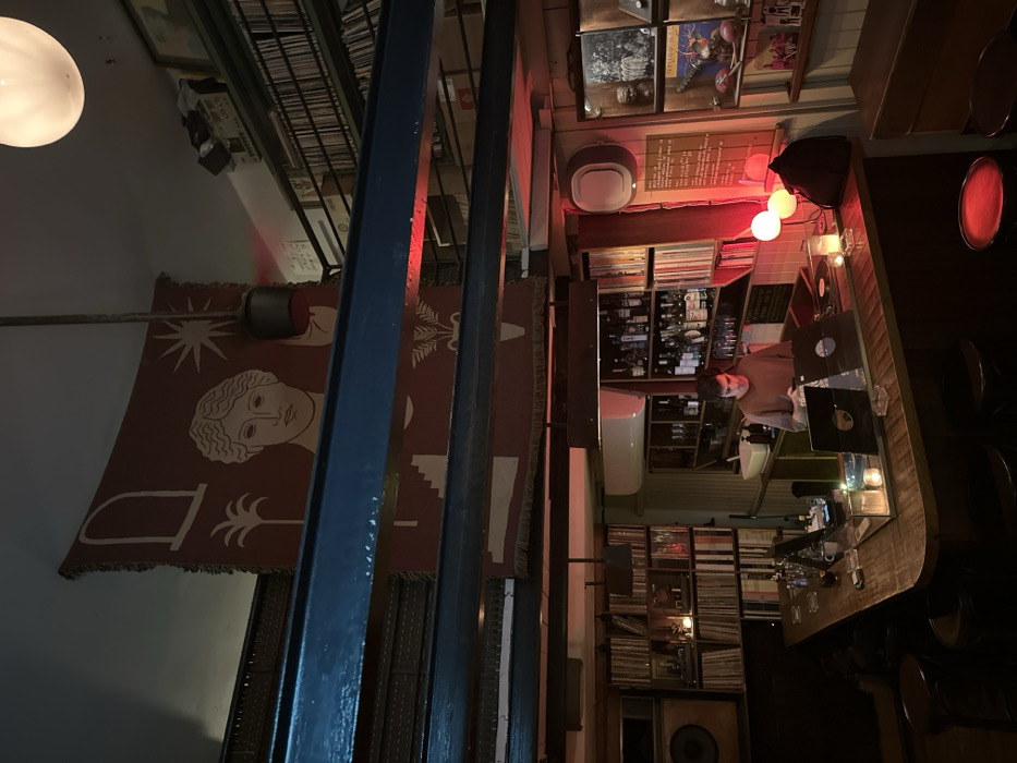This image captures the interior of Alfa Omega, a record bar situated in Tel-Aviv, Israel. The establishment is dimly lit with warm, amber, and red light sources.
The central feature is a curved, dark wood bar counter. Behind this counter, one male individual with short dark hair, wearing a light brown long-sleeved shirt, is observed looking down at a small handheld electronic device. On the bar counter in front of him are two turntables, each with a vinyl record in place. Several lit candles in glass holders and a modern desk lamp with two spherical red light sources are also on the counter.
Behind the bartender, shelves are stocked with bottles of liquor and glassware. A white utility sink is visible. The walls to the left and right of the bar are floor-to-ceiling bookshelves densely packed with vinyl records. On the far left, a large, light-colored speaker cabinet is positioned above the record shelves. On the right, shelves hold a mix of records and decorative objects, including a small bust, framed items, and what appears to be a pair of boxing gloves.
Above the main bar area, two prominent blue industrial I-beams span horizontally. A large, reddish-brown tapestry hangs from the ceiling, displaying stylized beige illustrations including a classical female face, a starburst, an archway, a palm tree, and an amphora with wheat stalks. Above the right side of the bar, a mezzanine level, also lined with records, is visible. A single white spherical pendant light hangs from the ceiling in the upper right quadrant of the image. Dark-colored bar stools are visible in the foreground, suggesting patron seating.