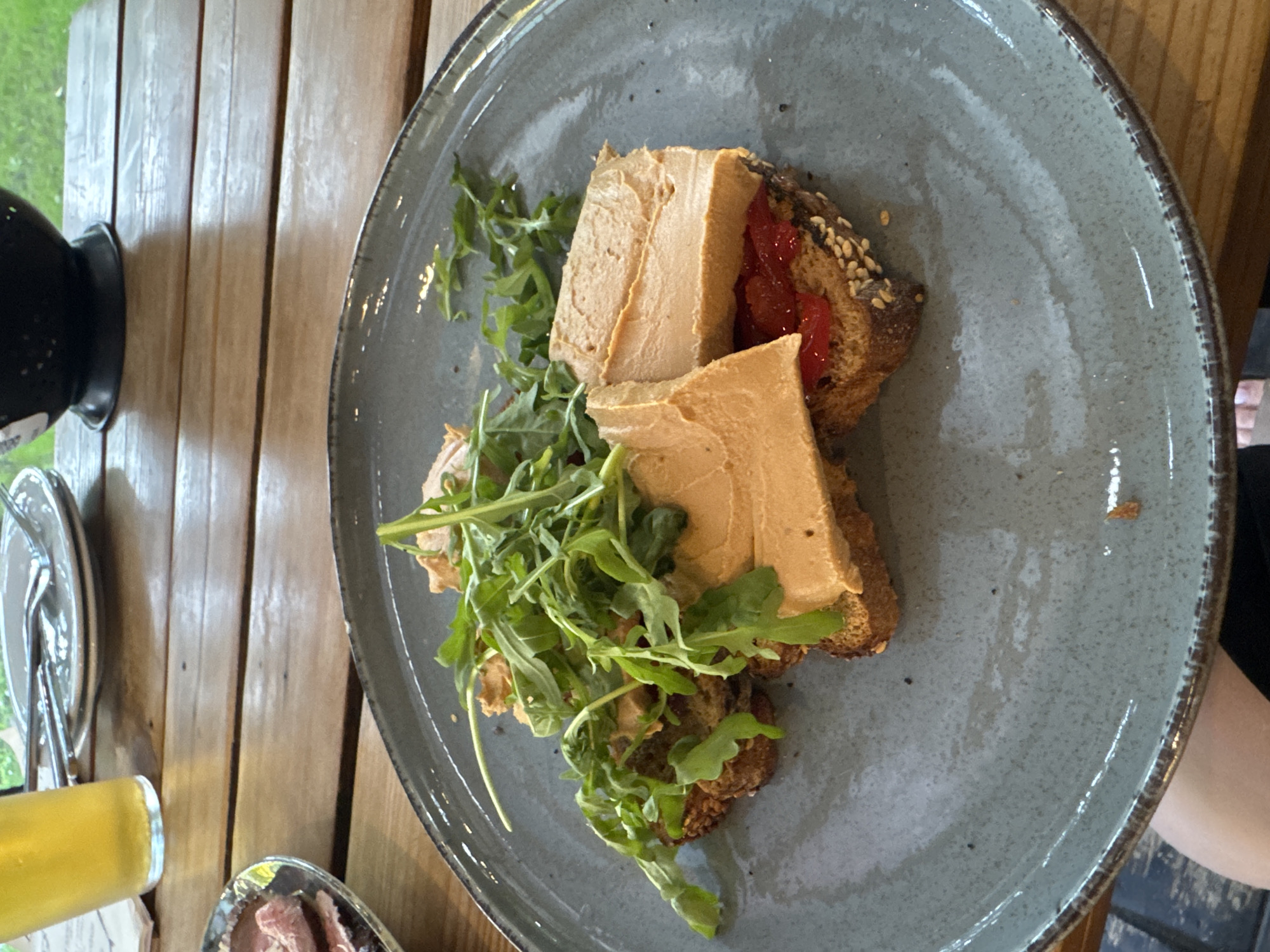 A close-up, eye-level shot shows a plate of pâté toast served with arugula and a glass of beer on a wooden table. The pâté is a smooth, light brown spread, served in thick slices. It is layered on top of toast, which appears to be seeded rye bread. Red pepper jam is visible beneath the pâté on one of the toast slices. A generous portion of fresh arugula is piled on top of the pâté. To the left of the plate, a glass of golden beer is visible. The table is made of weathered wooden planks. In the background, other tableware, a dark bowl, and greenery can be seen. This image was taken in Kremenchuk, Ukraine.