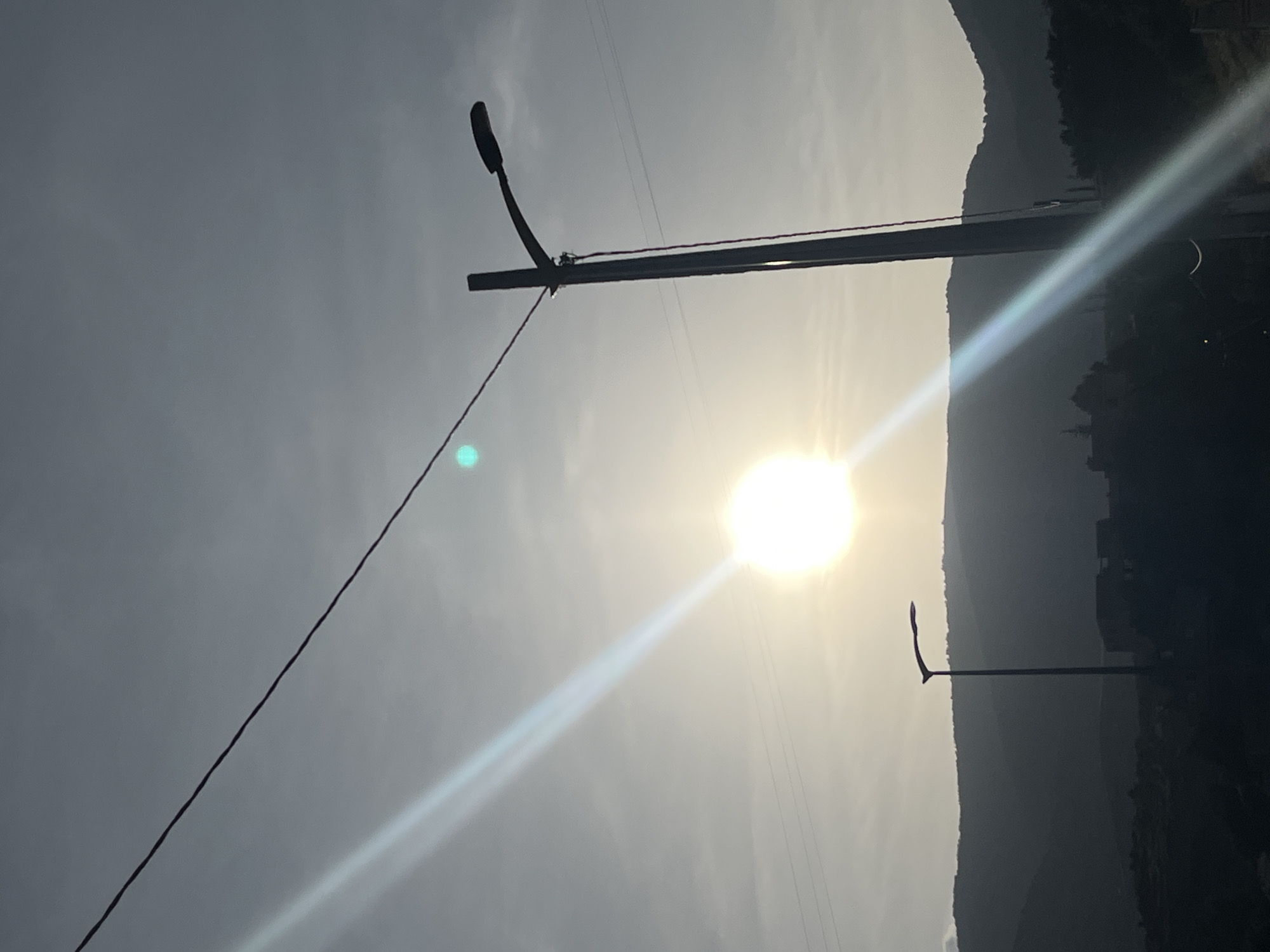 The image depicts a landscape in Ouaouizert, Morocco, possibly during sunrise or sunset, given the low angle of the sun. The sun is obscured by the light, however, there are a set of hills to the right. Two power poles are visible, one in the foreground and one in the background. A power line stretches across the top left of the image. The sky is overcast. The image is shot at an angle.