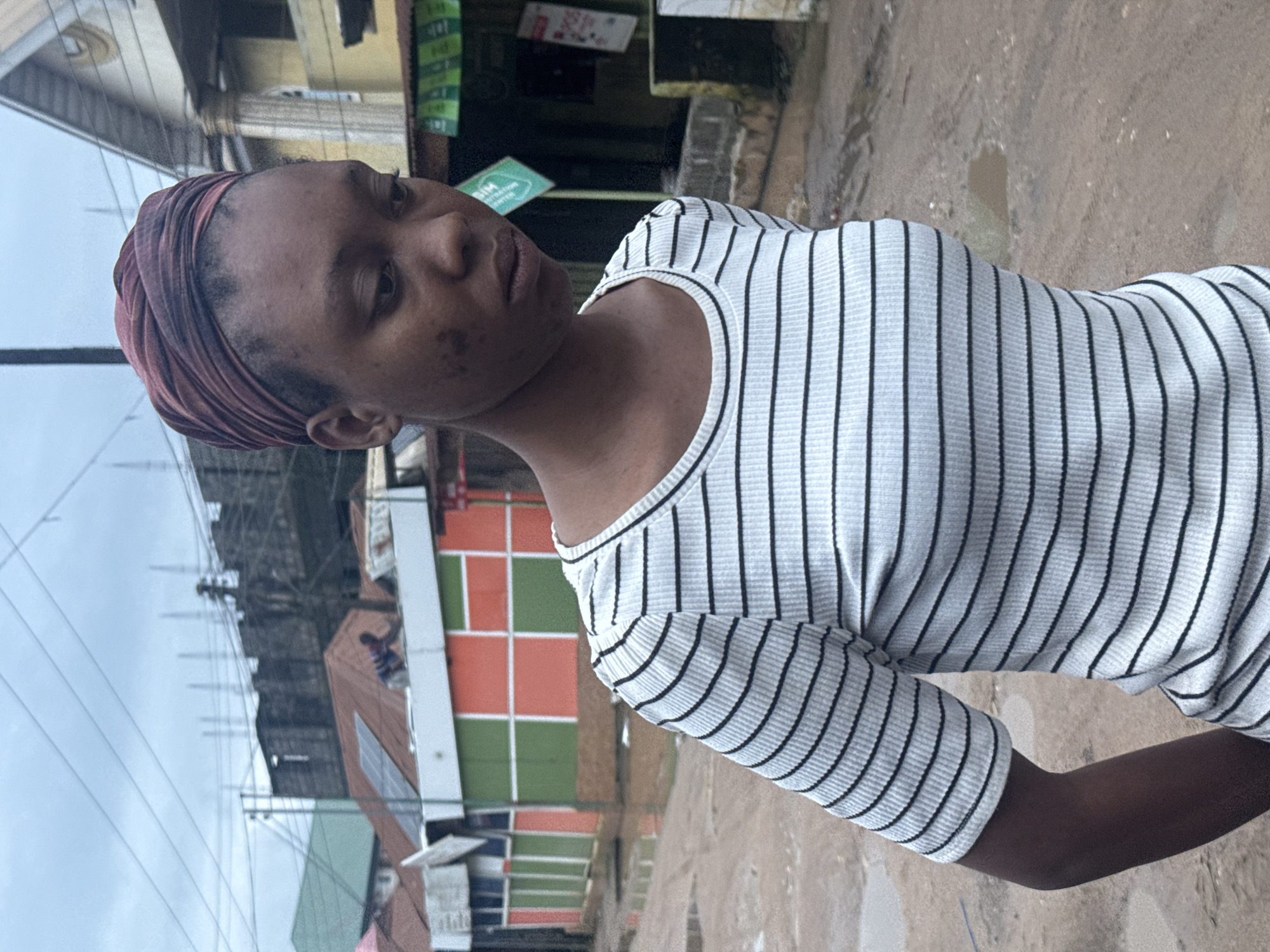 A young woman with dark skin and dark hair is standing outdoors, looking down and to her right. She is wearing a maroon headwrap and a white and black striped t-shirt. Her face has some dark spots. In the background, there are buildings, some under construction, and a muddy, wet street with puddles. The sky is overcast. The scene appears to be a street in Ihiagwa, Nigeria, with evidence of recent rain.