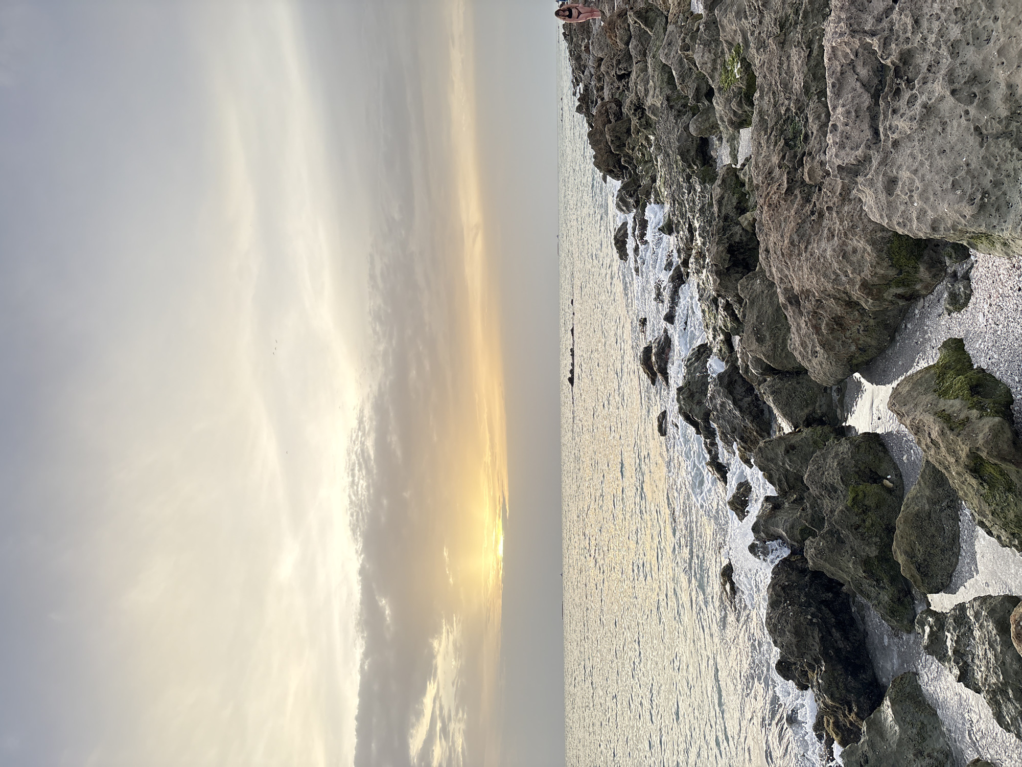 A wide shot captures a rocky shoreline under a cloudy sky with the sun setting over the ocean. The sun's rays create a golden glow on the horizon, breaking through the overcast clouds. The foreground is dominated by large, textured rocks, some covered in moss, with waves gently crashing against them and pooling in the crevices. A person wearing a dark bikini stands on the rocks on the right side of the image, facing the ocean. The water is a calm, shimmering expanse, reflecting the soft light of the sunset. In the distance, a small boat is visible on the water, and faint landmasses or structures are discernible on the horizon. The overall scene evokes a serene, end-of-day atmosphere. This image is of a beach in Venice, Florida, USA.