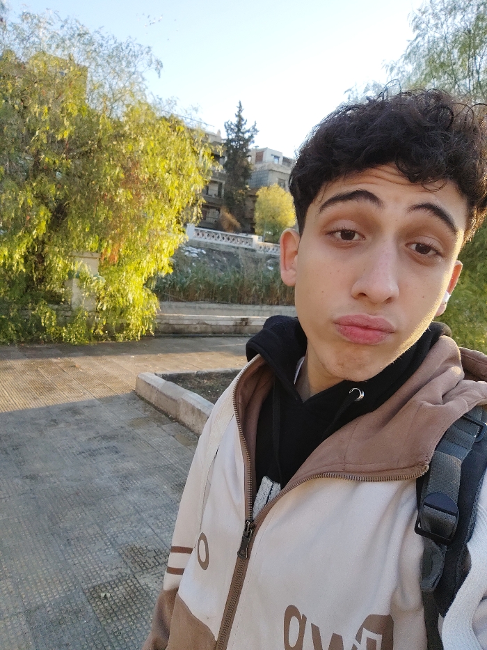 The image captures a close-up outdoor portrait of a young male, appearing to be in his late teens or early twenties, within As-Salihiya Municipality, Syria. He has dark, curly hair and light brown eyes, looking directly at the camera with a slight pout. He is dressed in a black hooded sweatshirt layered under an unzipped, two-tone jacket that is cream/off-white on the upper portion and light brown on the lower. Partial dark lettering is visible on the jacket. A dark-colored backpack with a gray strap is visible on his right shoulder, and he wears a small silver hoop earring in his left ear and a white earbud in his right ear.
The background features a paved ground surface with a low concrete barrier on the right side. Behind the subject, a line of trees with green and yellowish foliage is visible, illuminated by what appears to be morning or late afternoon sunlight, casting long shadows. Further back, a stone retaining wall topped with a white decorative balustrade is present, with more vegetation below it. Beyond the wall, multi-story buildings and a tall, dark coniferous tree are visible on an elevated terrain against a clear, pale blue sky.