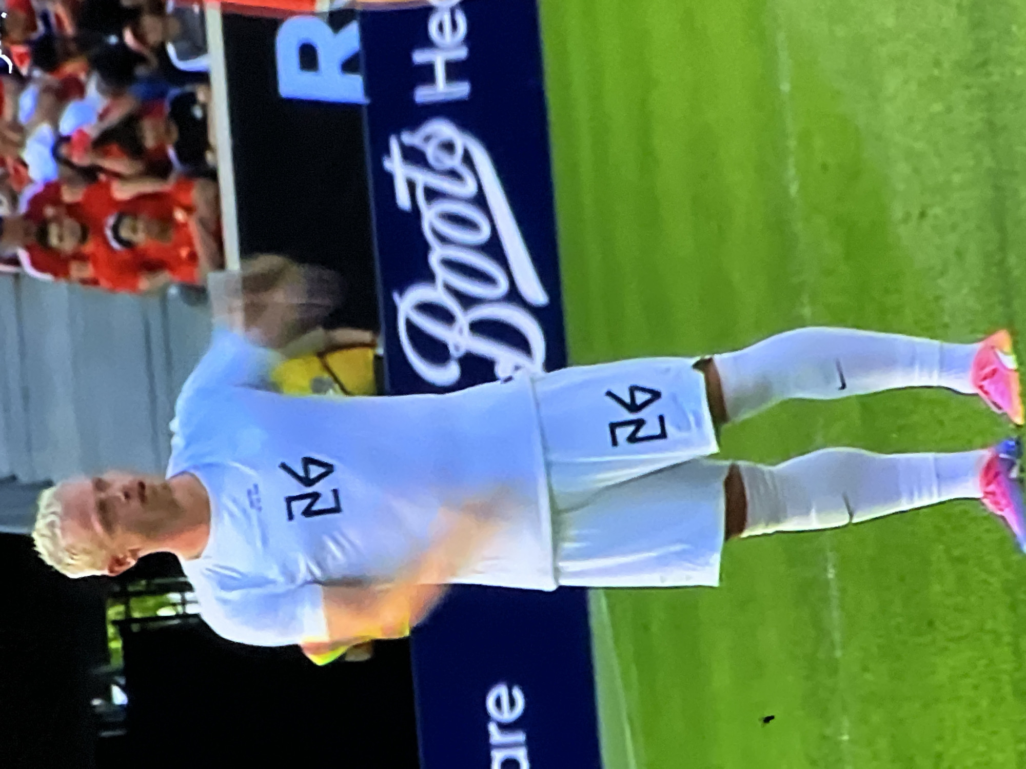 The image depicts a male soccer player on a green grass field within an outdoor sports stadium, specifically located in the city of Casablanca, Morocco. The player, with short blonde hair and fair skin, is positioned centrally, wearing a white short-sleeved athletic shirt and matching white shorts, both prominently displaying the stylized black number "26". He wears white athletic socks with a black Nike swoosh visible on his left leg, and multi-colored (pink and blue/purple) soccer cleats. His right arm is partially blurred, suggesting motion. In the background, seated spectators are visible in stadium stands, predominantly wearing red attire. Below the spectators, a dark blue advertising board is visible, featuring white text including "are", the prominent "Boots" logo in cursive script, and partially visible "He" (likely part of "Health") and a large "R". The scene is brightly lit.