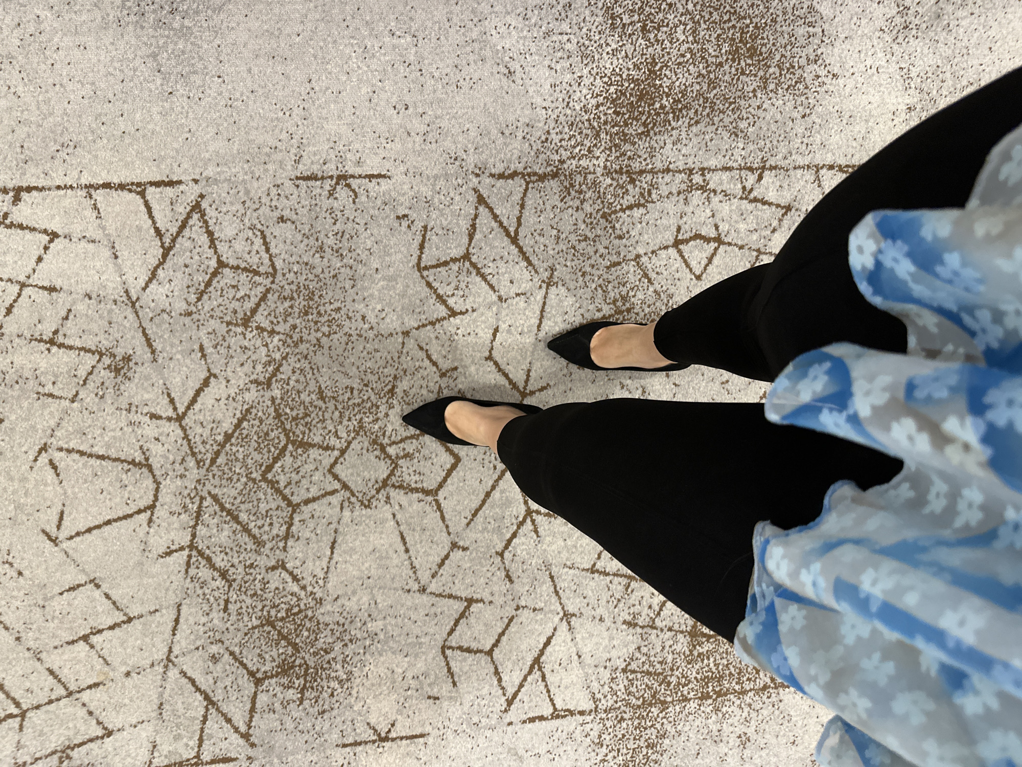 A high-angle shot shows a person standing on a carpet with a geometric pattern. The person is wearing black pants and black pointed-toe shoes. A portion of a light blue and white floral-patterned garment is visible. The location is not discernible from the image.
