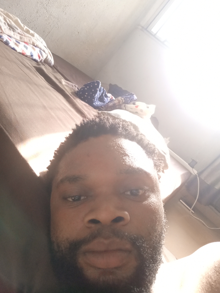 An adult male with dark skin, short dark hair, and a full beard is depicted in a close-up selfie, lying down in a bedroom located in Magboro, Nigeria. He is looking directly at the camera with a neutral expression. The upper right portion of his face is brightly illuminated. Behind him, to the left, is a bed with dark brown sheets, and a patterned blanket or pillow is visible near the head of the bed. Several folded or crumpled clothing items, including a blue item with white polka dots, are on the bed to the right of the blanket. The background features a light-colored wall with a window or light source on the upper right, which is overexposed, casting bright light into the room. A textured, light-colored ceiling is visible in the upper left. A white rectangular object and a descending white cable are faintly visible on the wall beneath the bright light source.